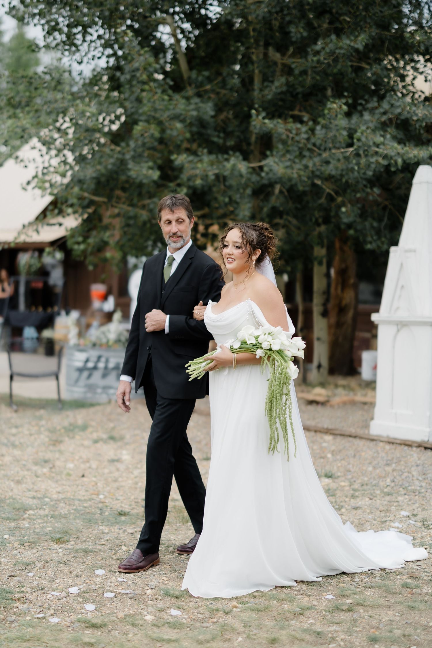 Bride in white off-shoulder gown holding bouquet walks arm-in-arm with groom in black suit at outdoor wedding venue.