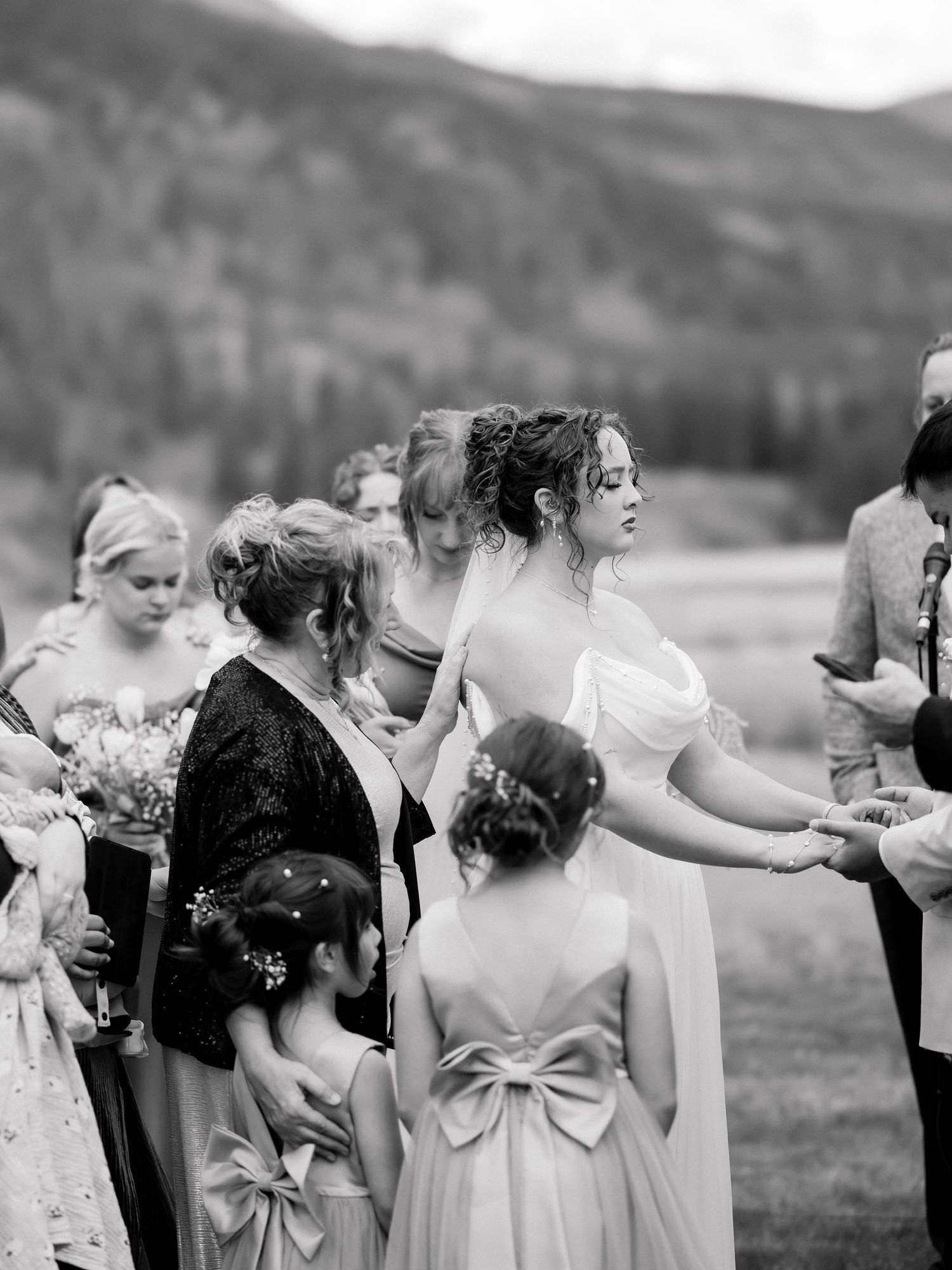 Black and white photo of bride in off-shoulder gown holding hands with wedding party in mountains.