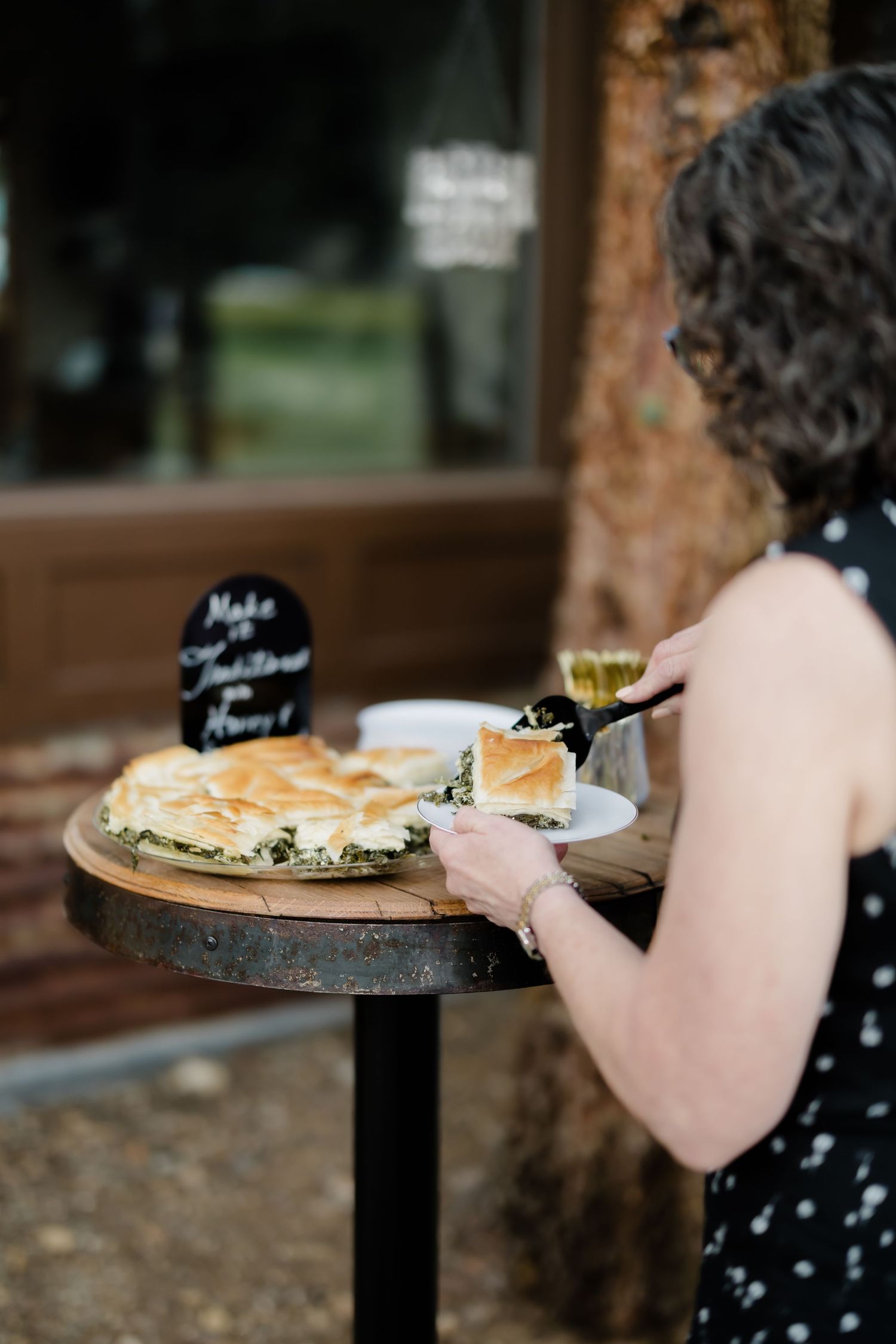 Woman serving spinach pie at an outdoor event table with a chalkboard sign and wine glass nearby.