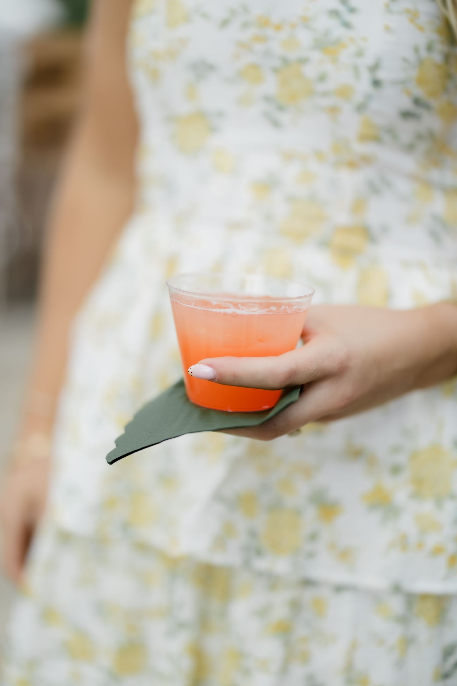 Woman in floral dress holding a clear plastic cup with orange cocktail and a green leaf garnish.