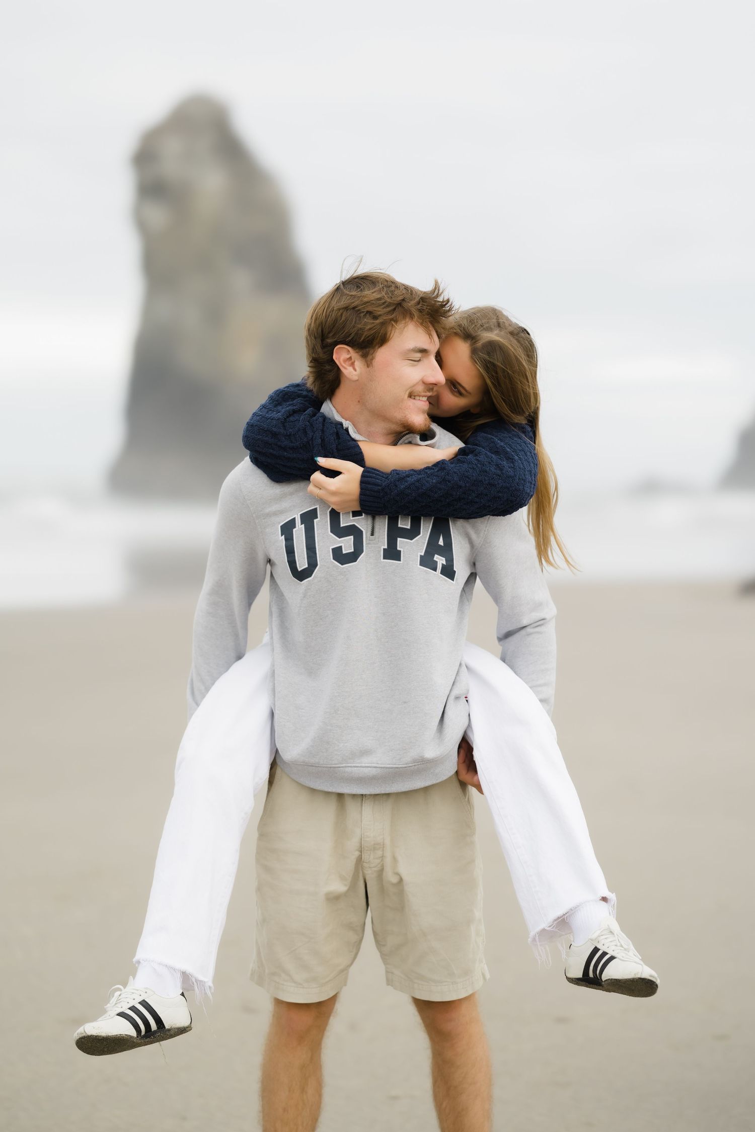 Man in USPA sweatshirt giving woman a piggyback ride on a sandy beach with dramatic rock formations behind them.