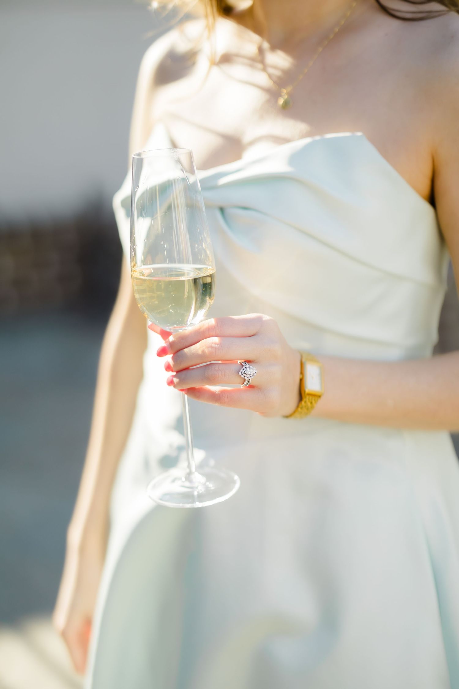 Bride in strapless gown holds champagne flute showcasing diamond engagement ring and gold bracelet.