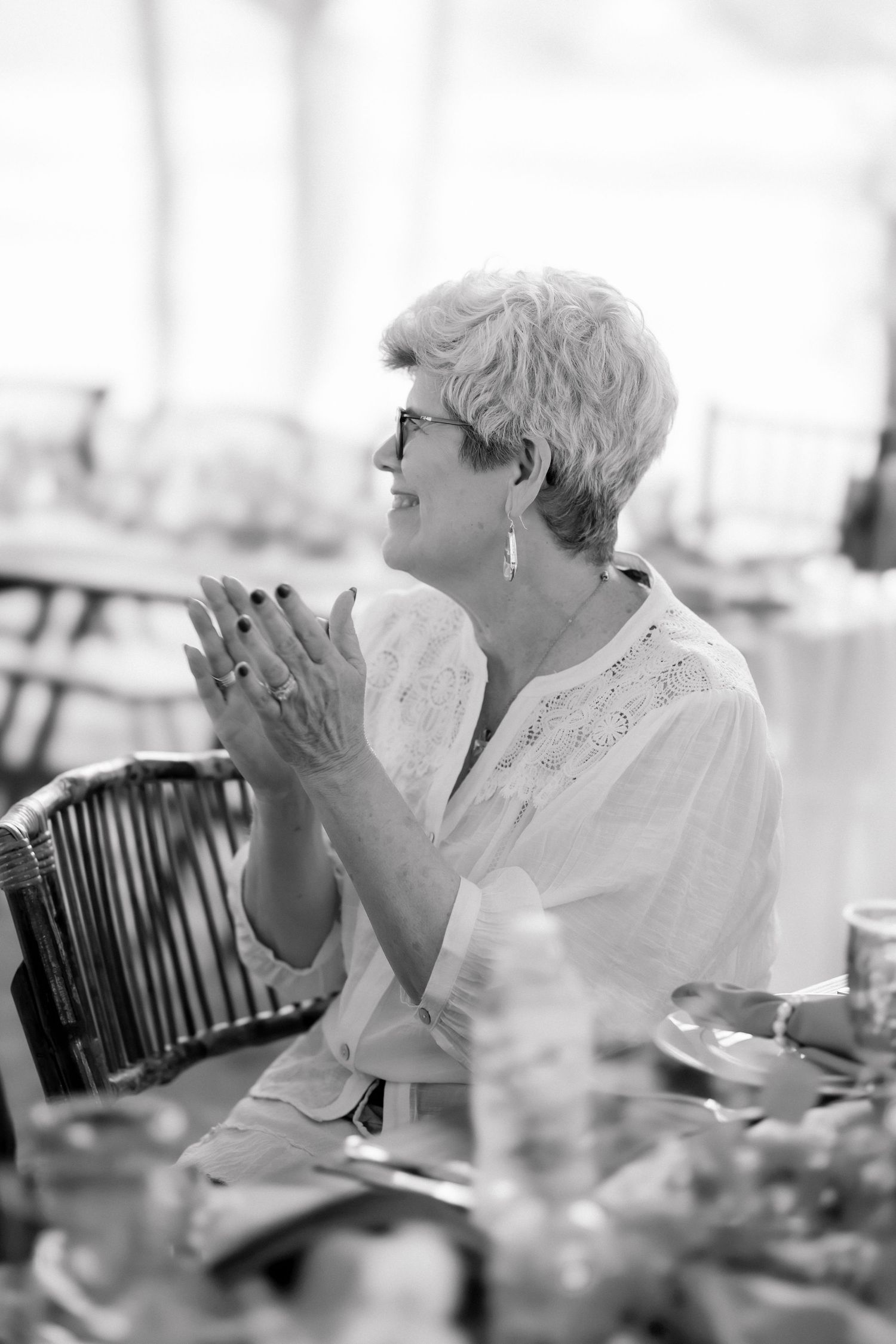 Older woman with short gray hair and glasses clapping at an outdoor event, black and white photo.