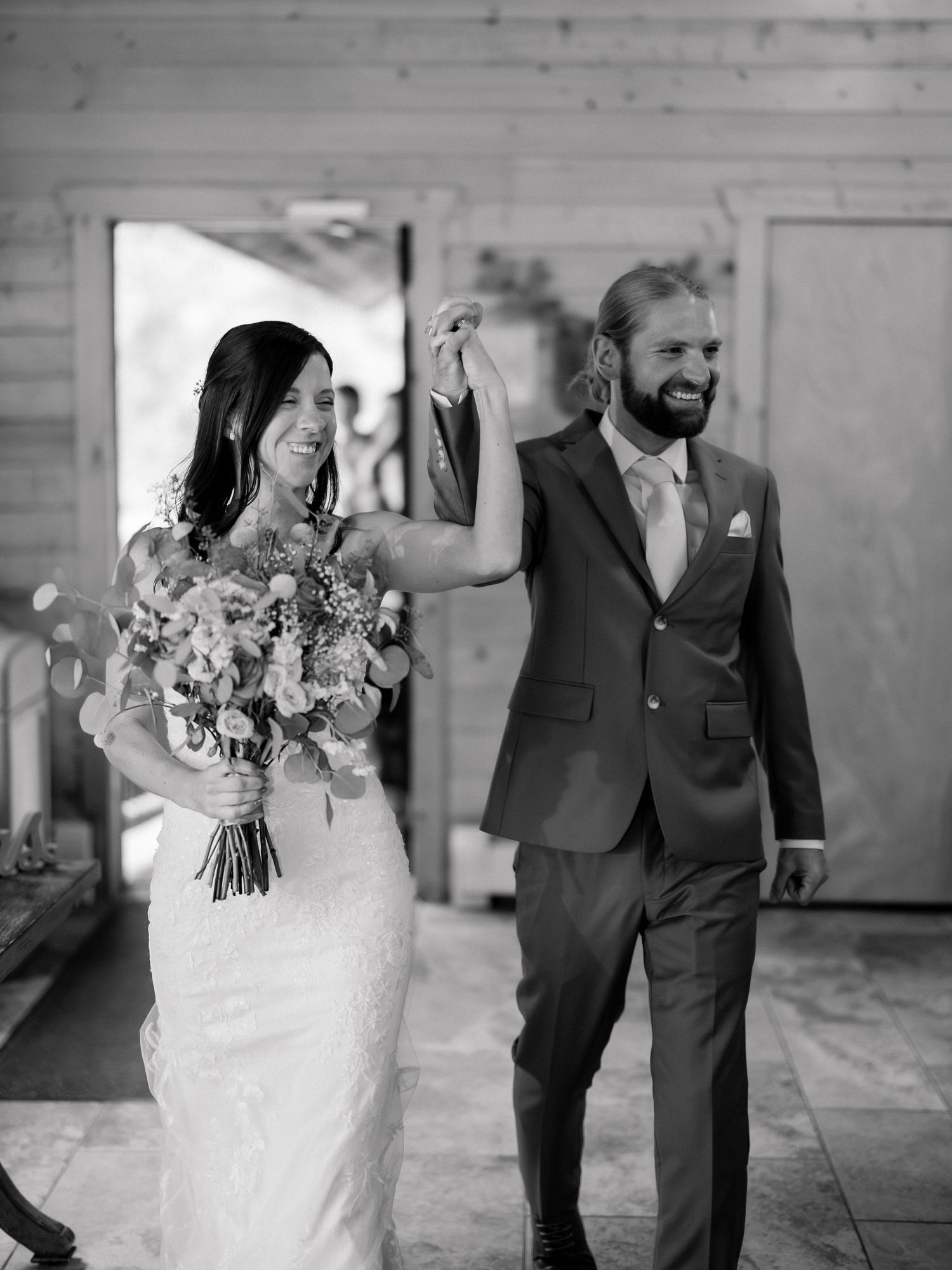 Black and white photo of a smiling bride holding a bouquet and groom in suit holding hands at their wedding reception.