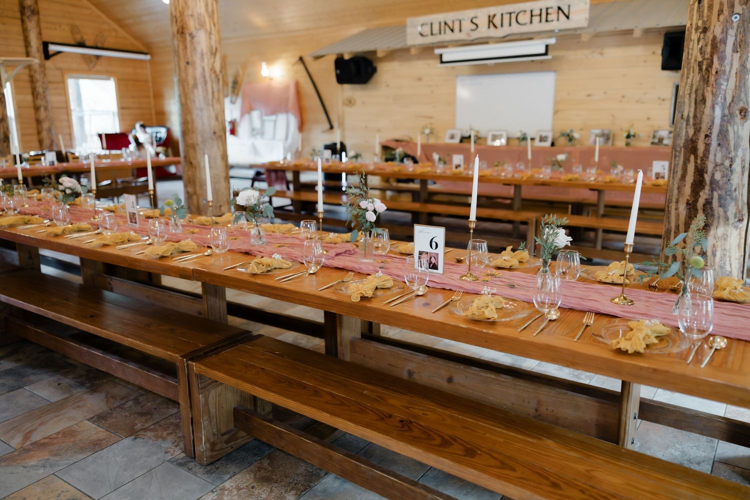 Rustic wedding reception hall with long wooden tables set with candles, pink runners, and floral centerpieces under Clint's Kitchen sign.