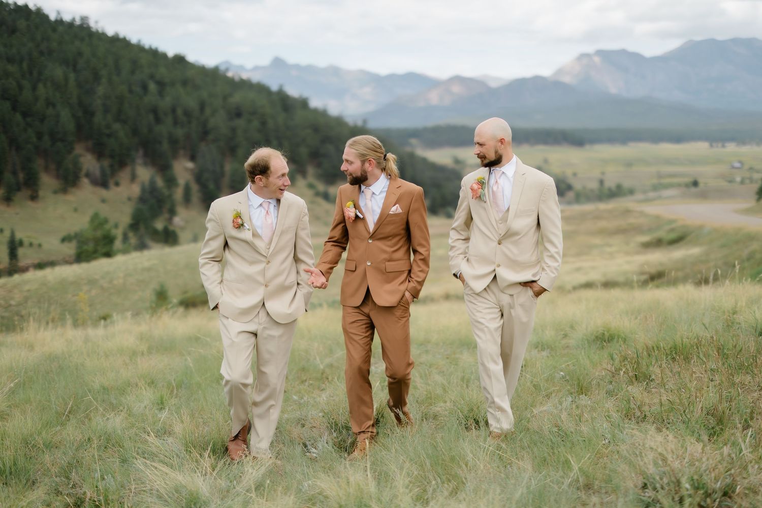 Groom in brown suit walks with two groomsmen in tan suits across a green mountain meadow.