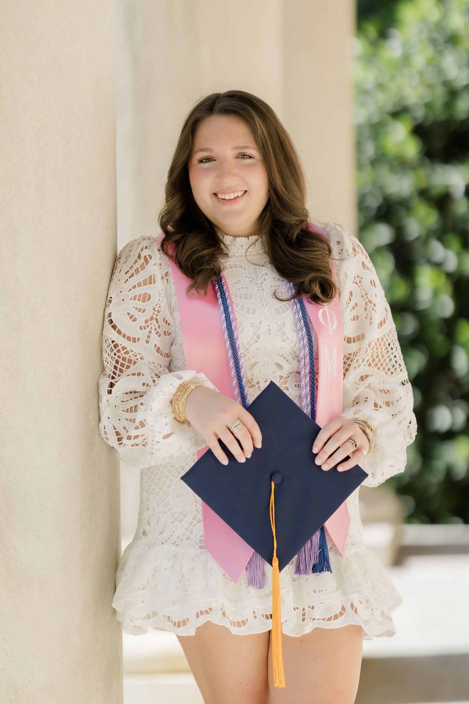 Smiling young woman in white lace dress holding navy graduation cap with gold tassel, wearing pink sash outdoors.