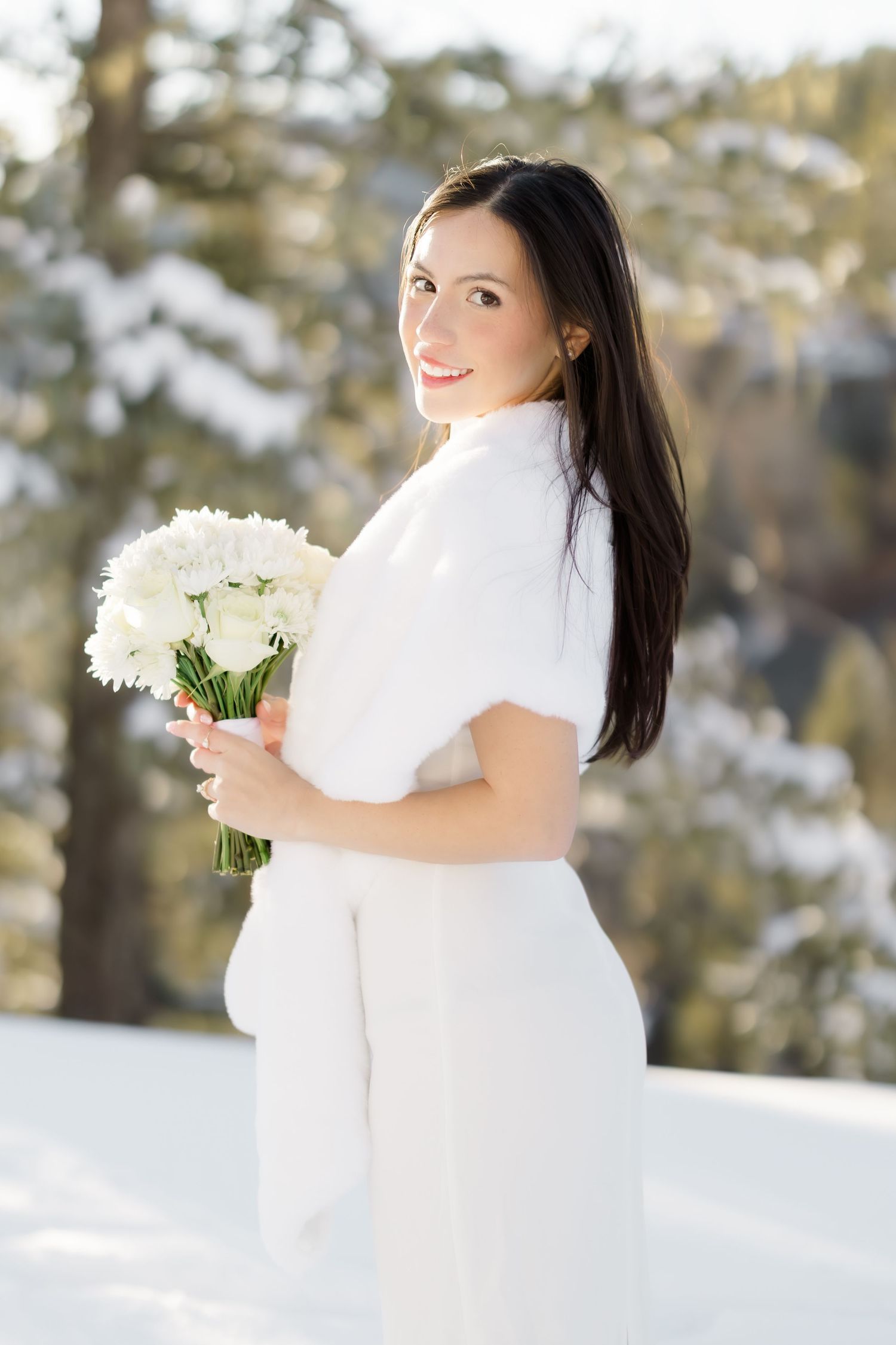 Bride in white dress and fur wrap holds white flower bouquet in snowy winter forest setting.