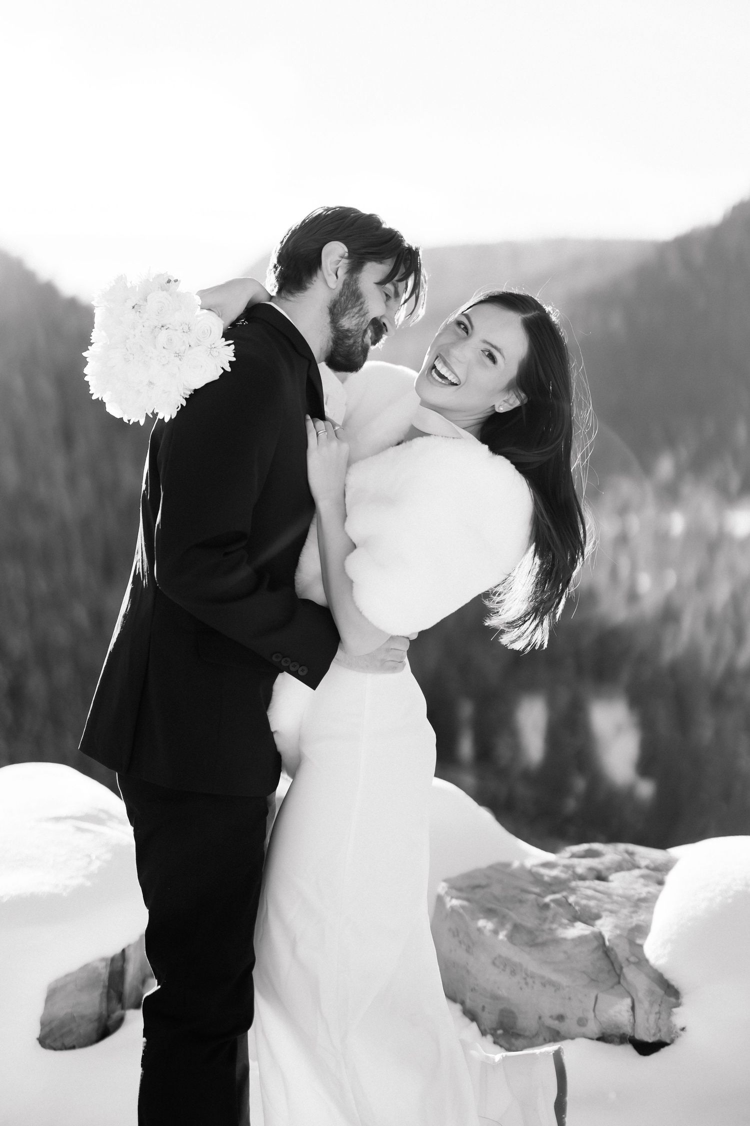 Black and white photo of laughing bride in fur stole embracing bearded groom holding white bouquet in snowy mountains.