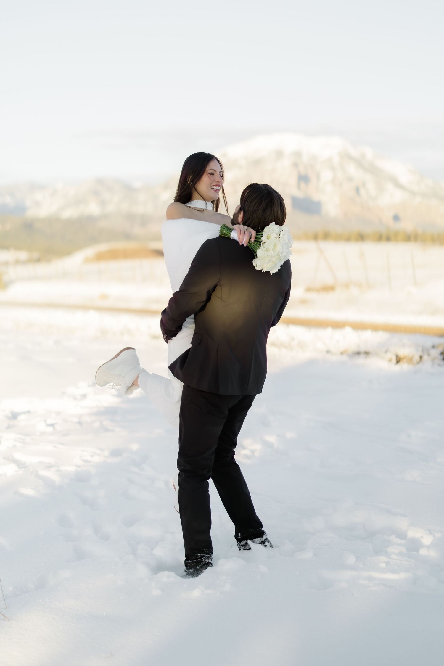 Groom lifts smiling bride in white wedding dress while standing in snowy mountain landscape.