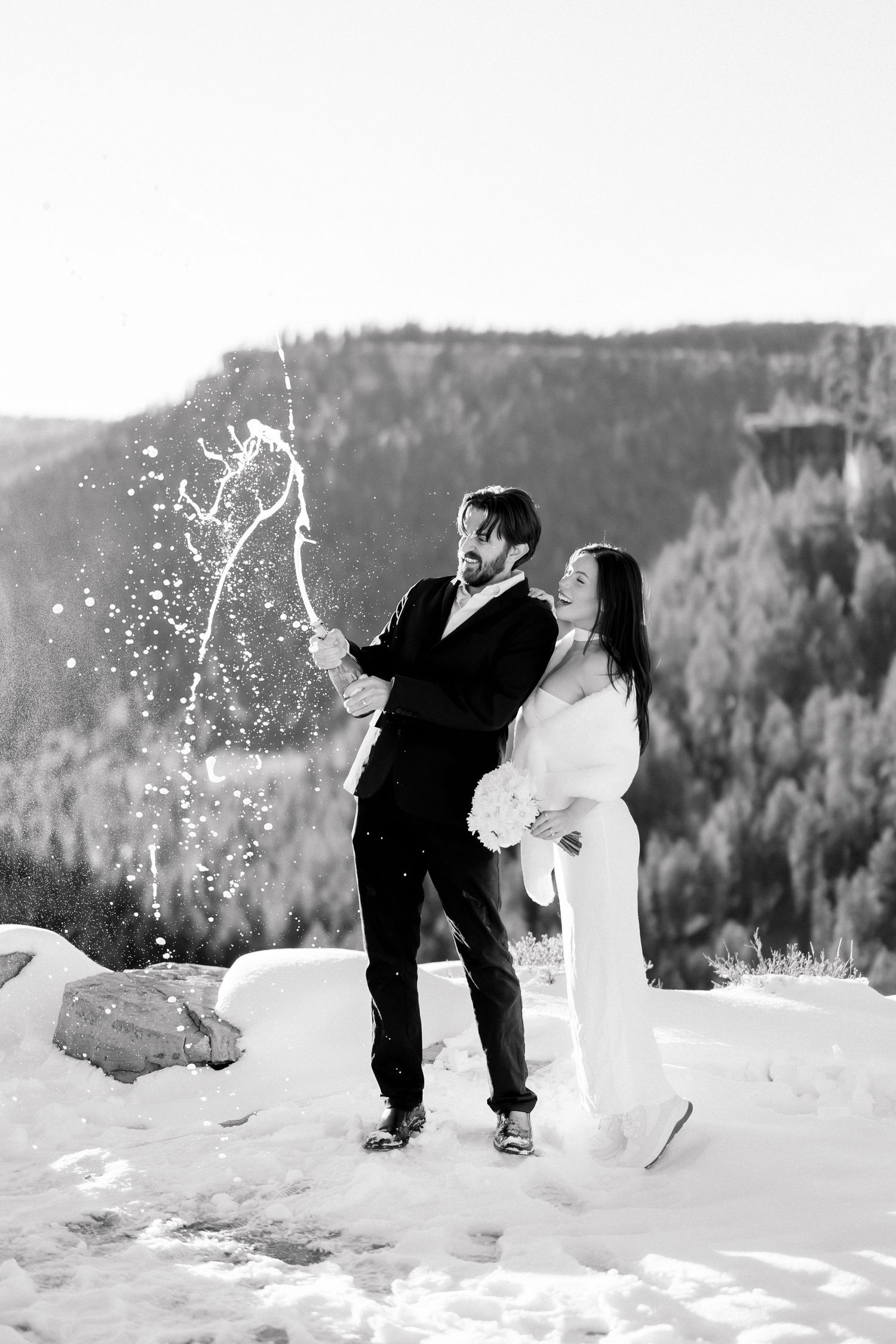 Black and white image of newlyweds celebrating with champagne spray in snowy mountain setting.