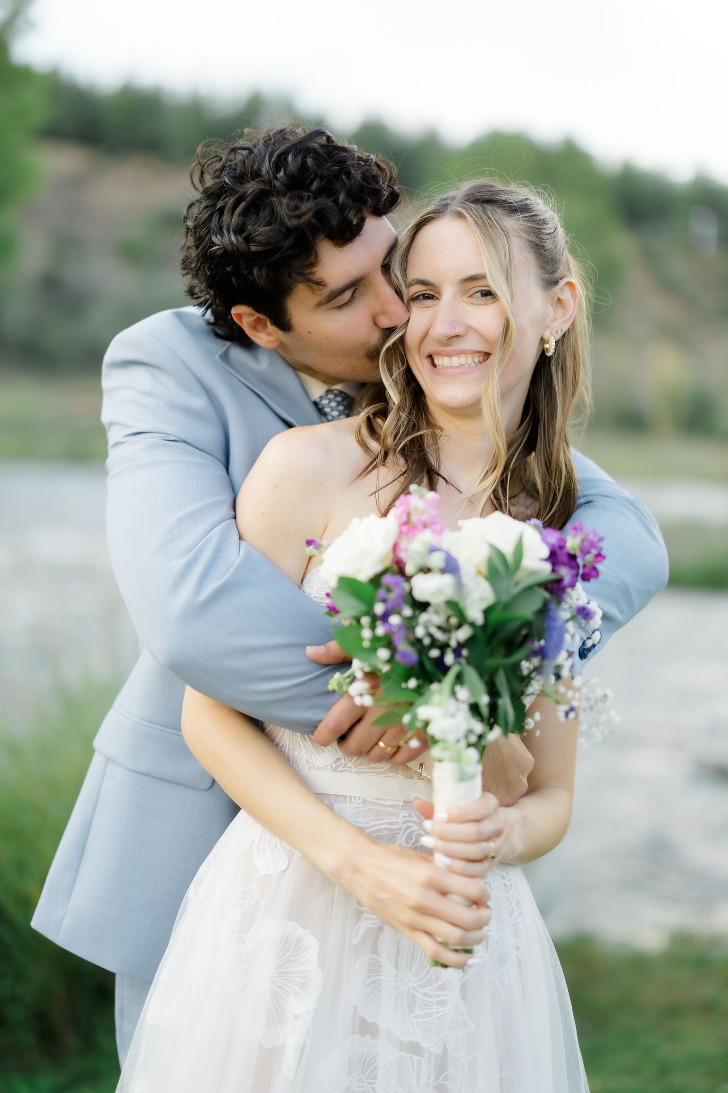 Groom in gray suit kisses bride's cheek as she smiles holding colorful wedding bouquet outdoors.