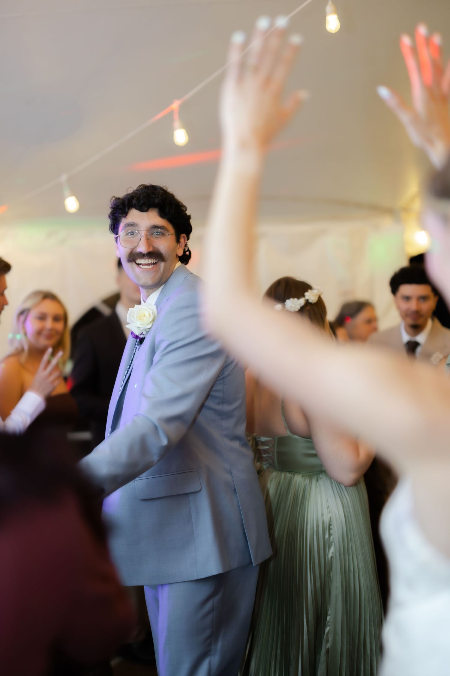 Smiling groom in gray suit with boutonniere dances with guests at a lively wedding reception under string lights.