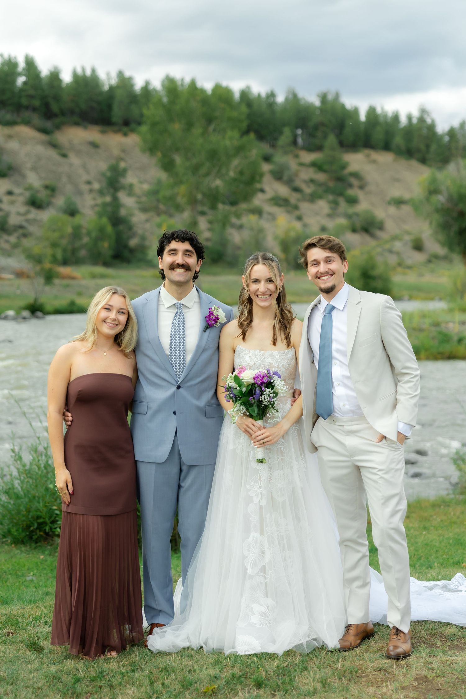 Wedding party of four posing outdoors by a river, bride in white gown holding bouquet, groom in gray suit.