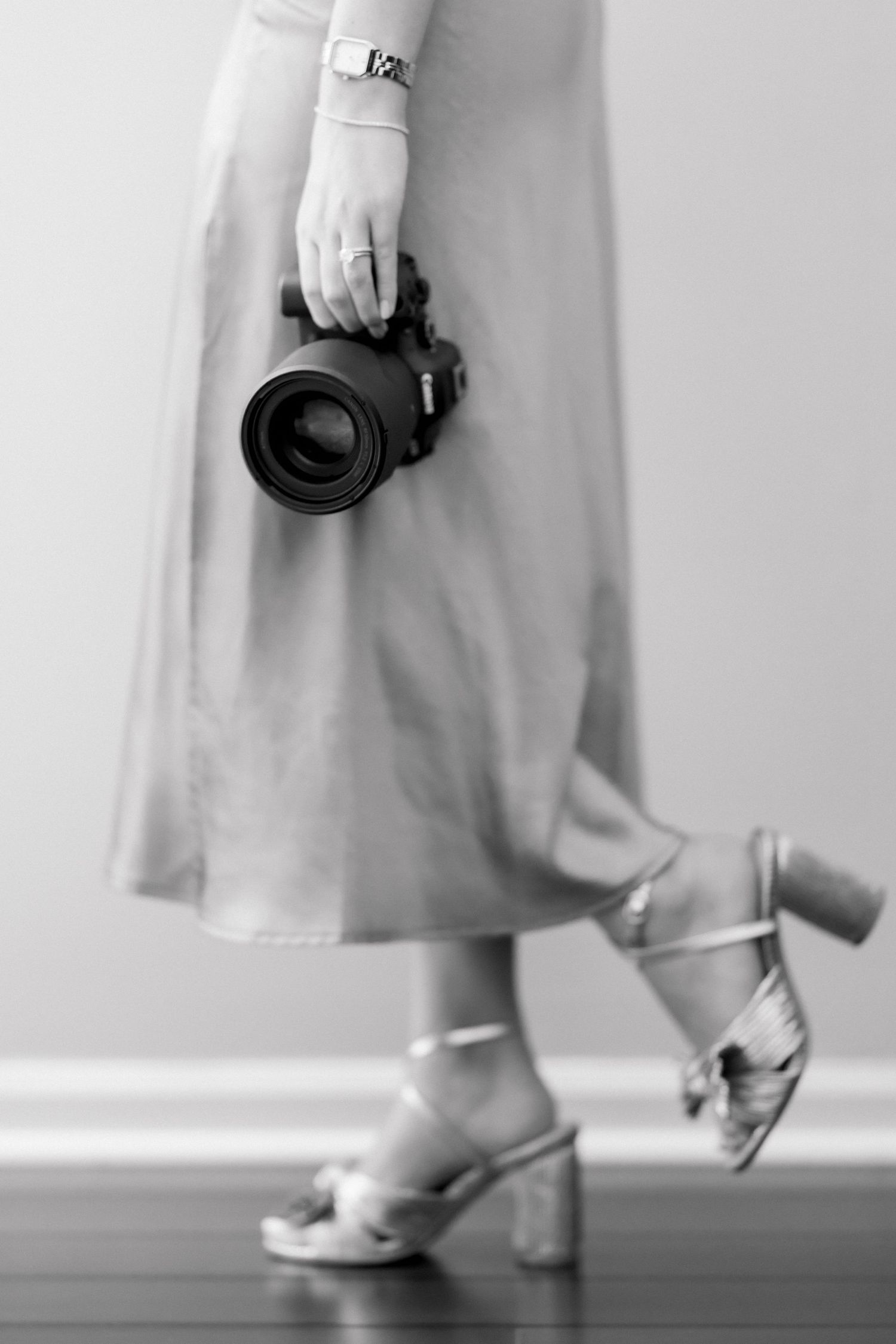 Black and white photo of a woman in a midi skirt and heeled sandals holding a DSLR camera at her side.