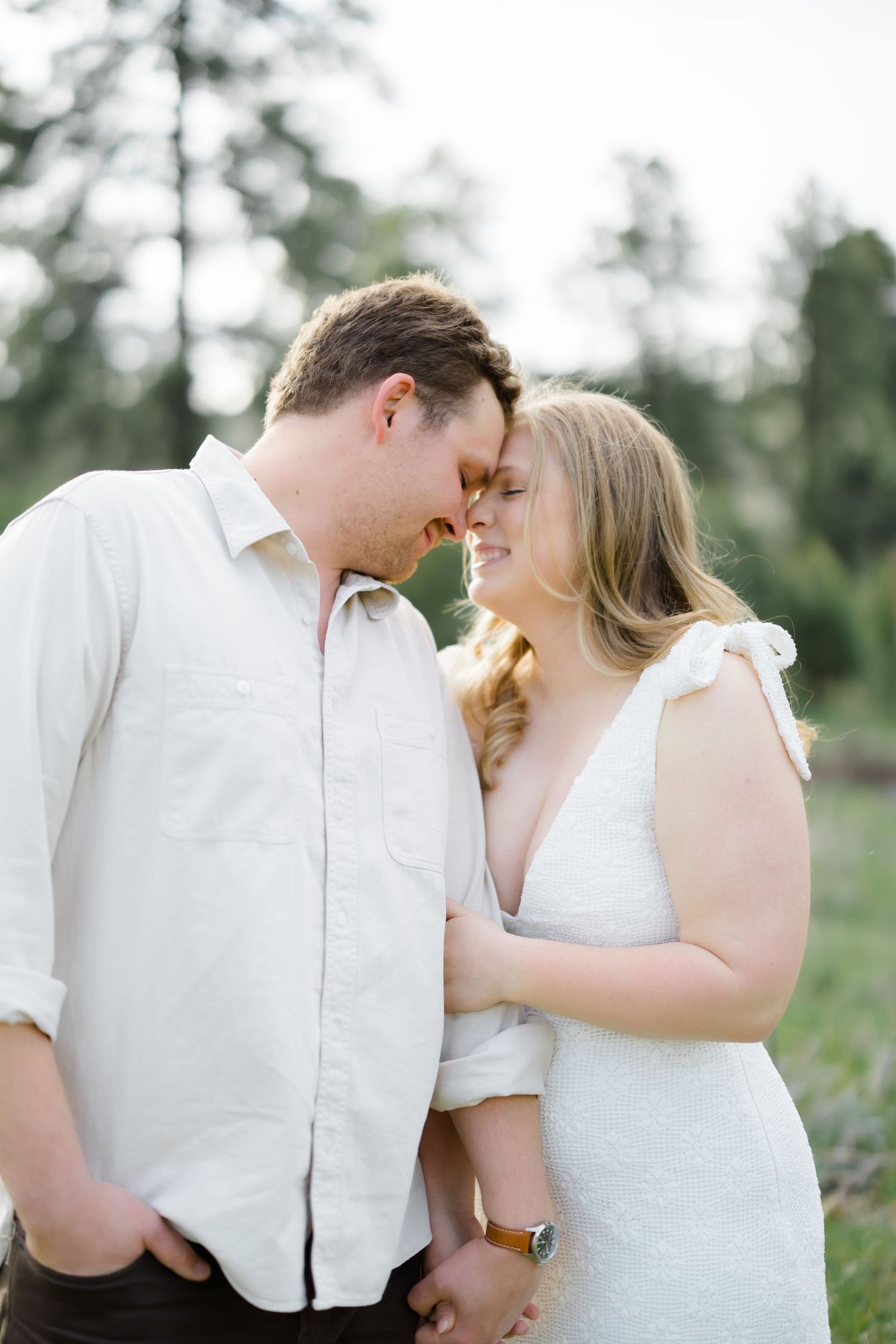 Couple in white outfits touching foreheads and smiling in a lush green outdoor setting.