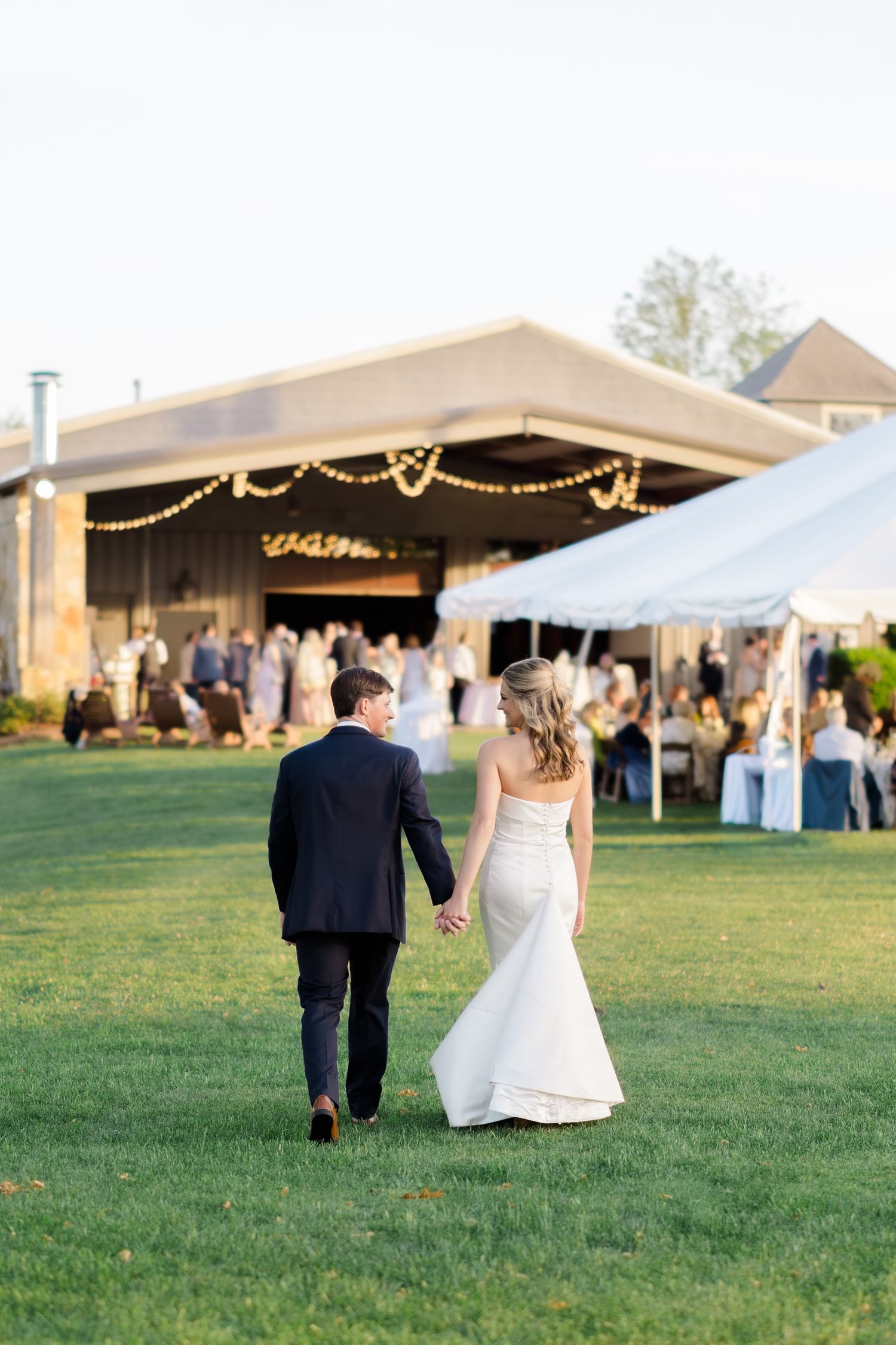 Bride and groom walk hand in hand across green lawn toward elegant tent reception with string lights at outdoor wedding.