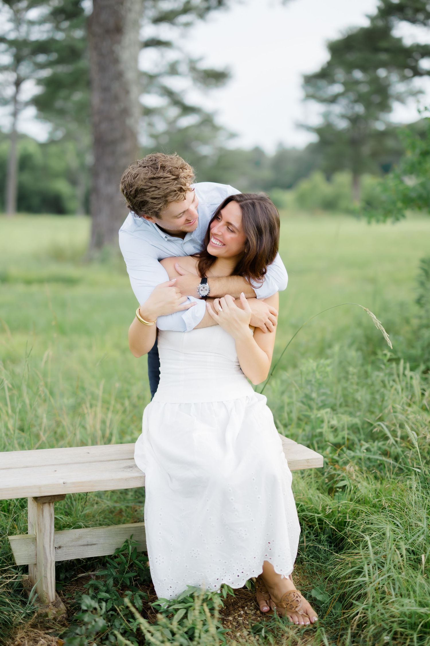 A couple shares a joyful embrace on a wooden bench in a green meadow, the man hugging the woman from behind.