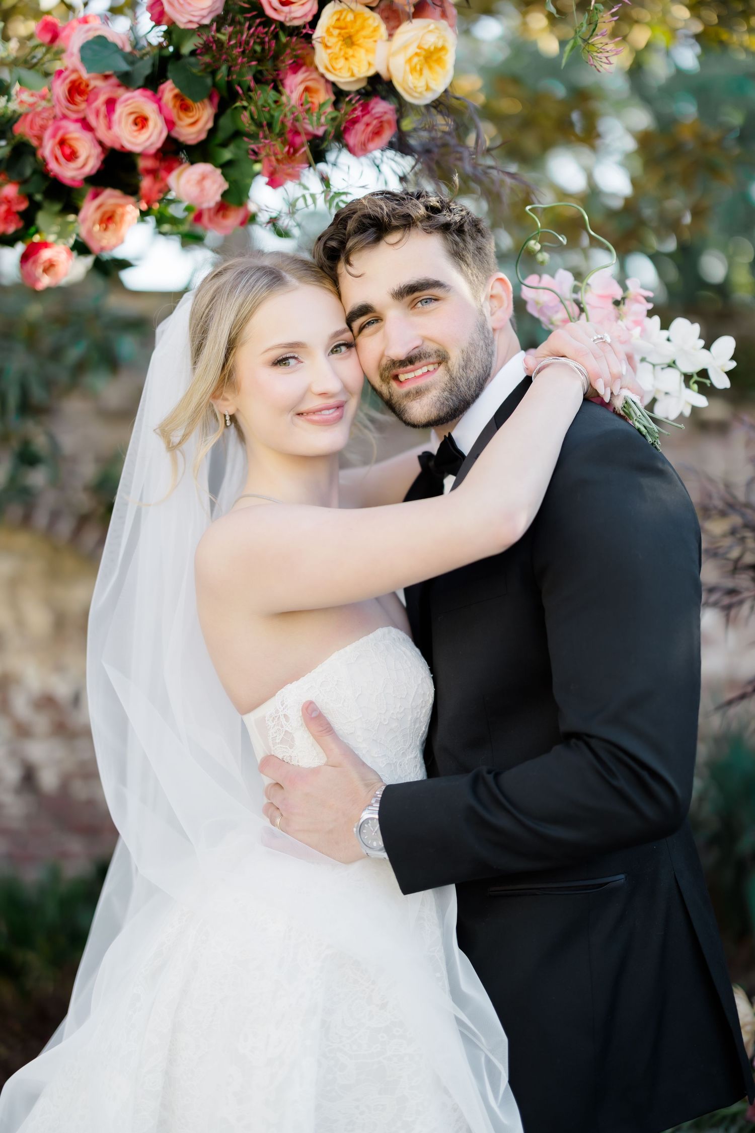 A romantic wedding portrait surrounded by coral and yellow floral arrangements in an outdoor garden.