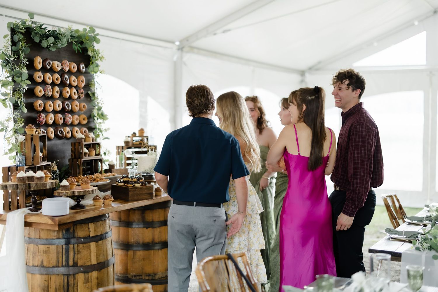 Guests mingle at an elegant outdoor wedding reception under a white tent with rustic barrel decor and donut wall.