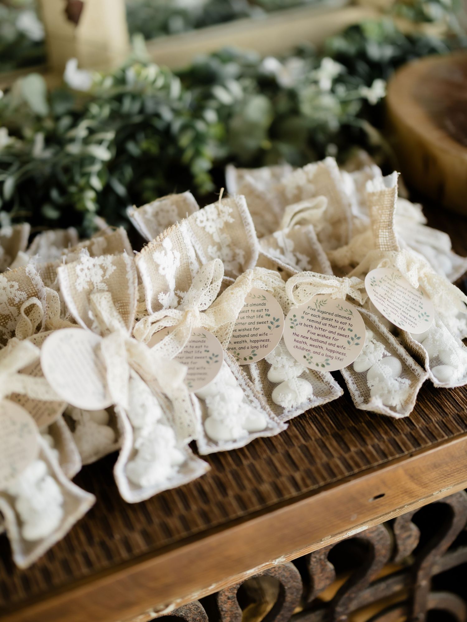 Rustic lace wedding favors with vintage tags displayed on a wooden table with greenery backdrop.