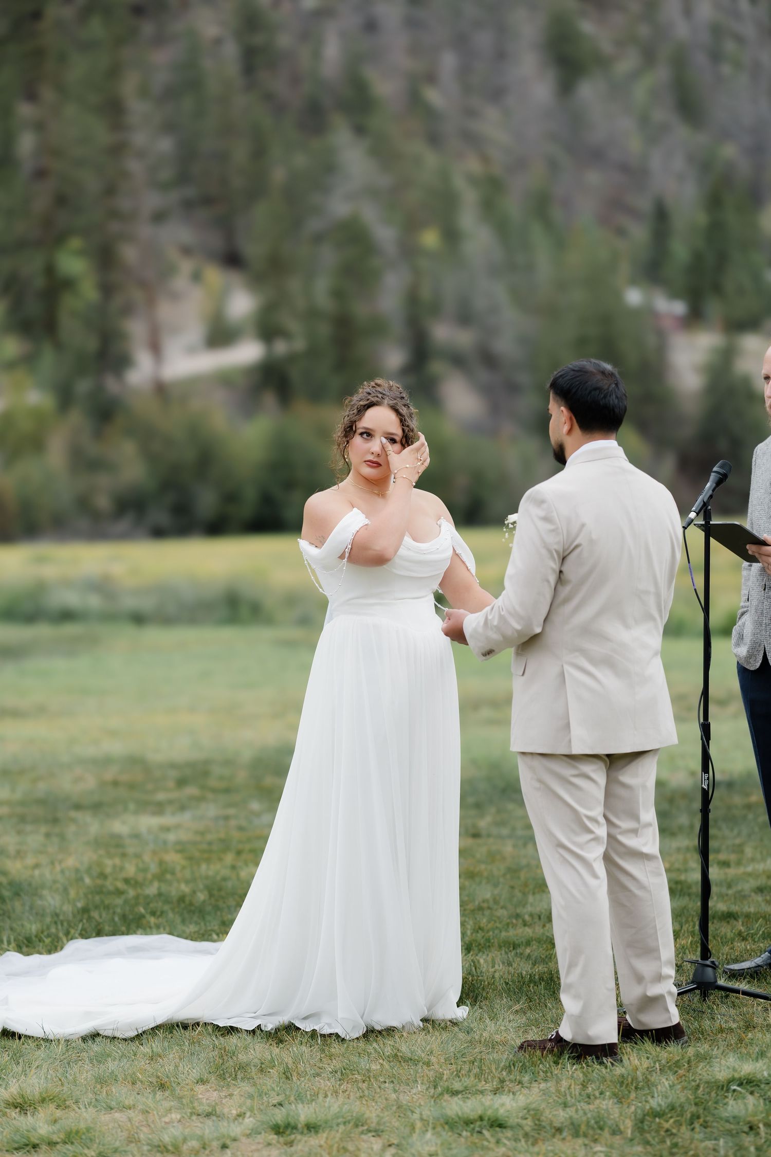 Emotional bride in off-shoulder white gown wiping tears during outdoor mountain wedding ceremony.