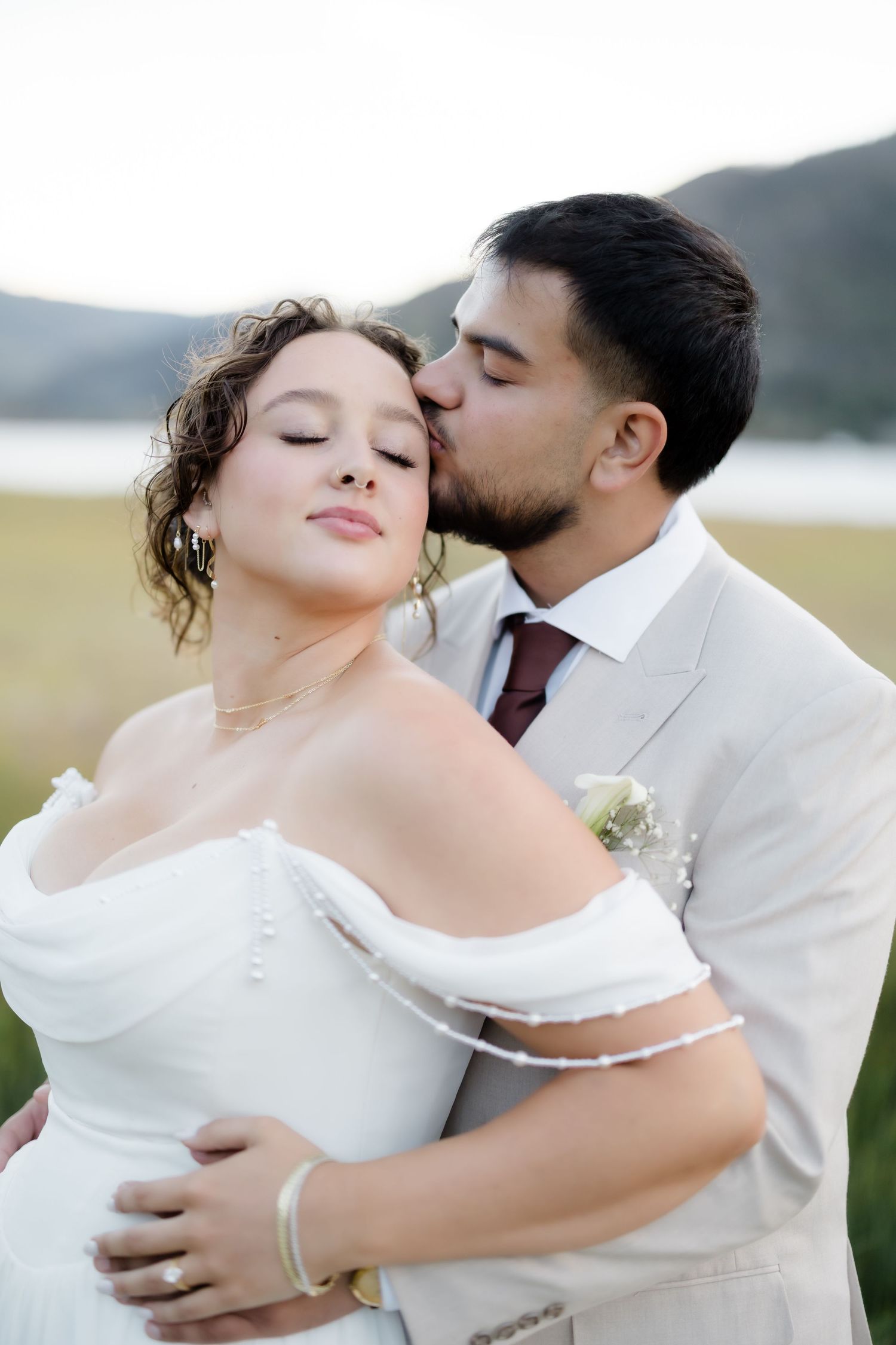 Groom kisses bride on cheek as she smiles with eyes closed, standing in a green field with mountains behind.