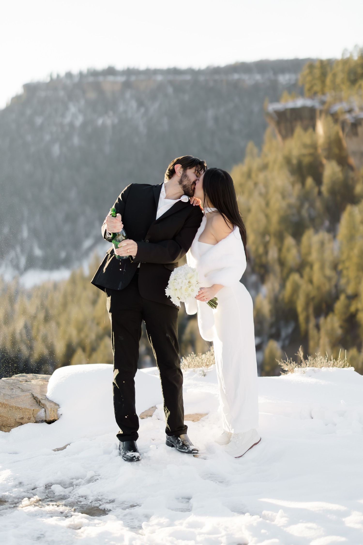 Newlyweds kissing while popping champagne bottle on a snowy mountain overlook with pine trees.