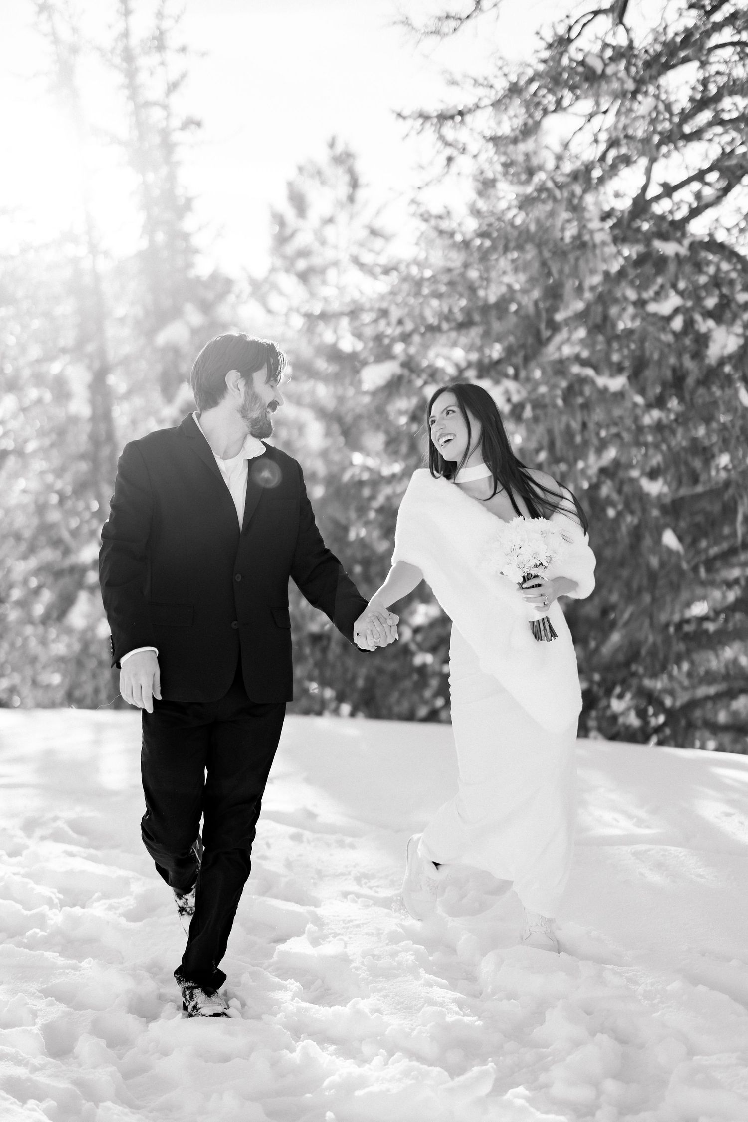 Black and white photo of smiling newlyweds holding hands walking through snowy pine forest.