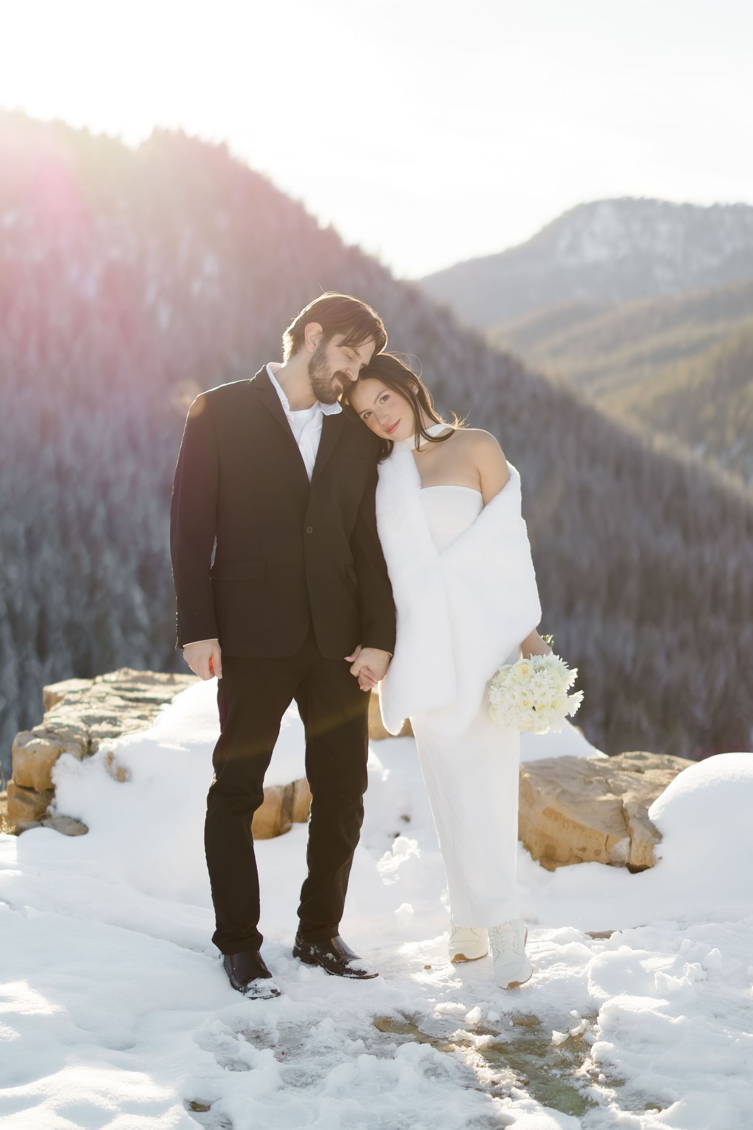 Bride and groom embracing in snowy mountain landscape, she holds white bouquet wearing white suit, he wears black tuxedo.