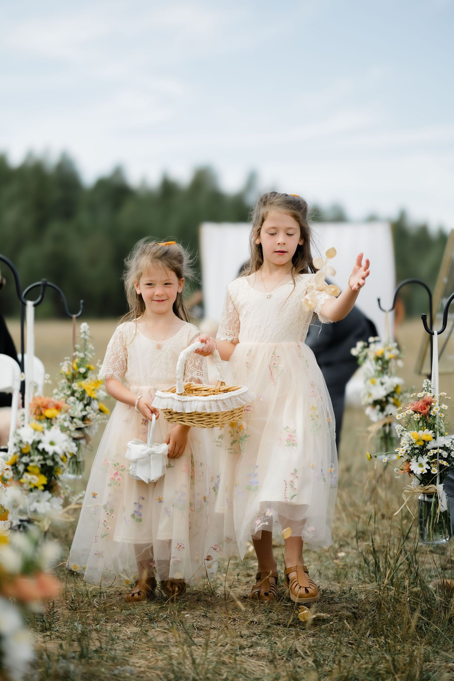 Two flower girls in floral dresses scatter petals at an outdoor wedding ceremony.