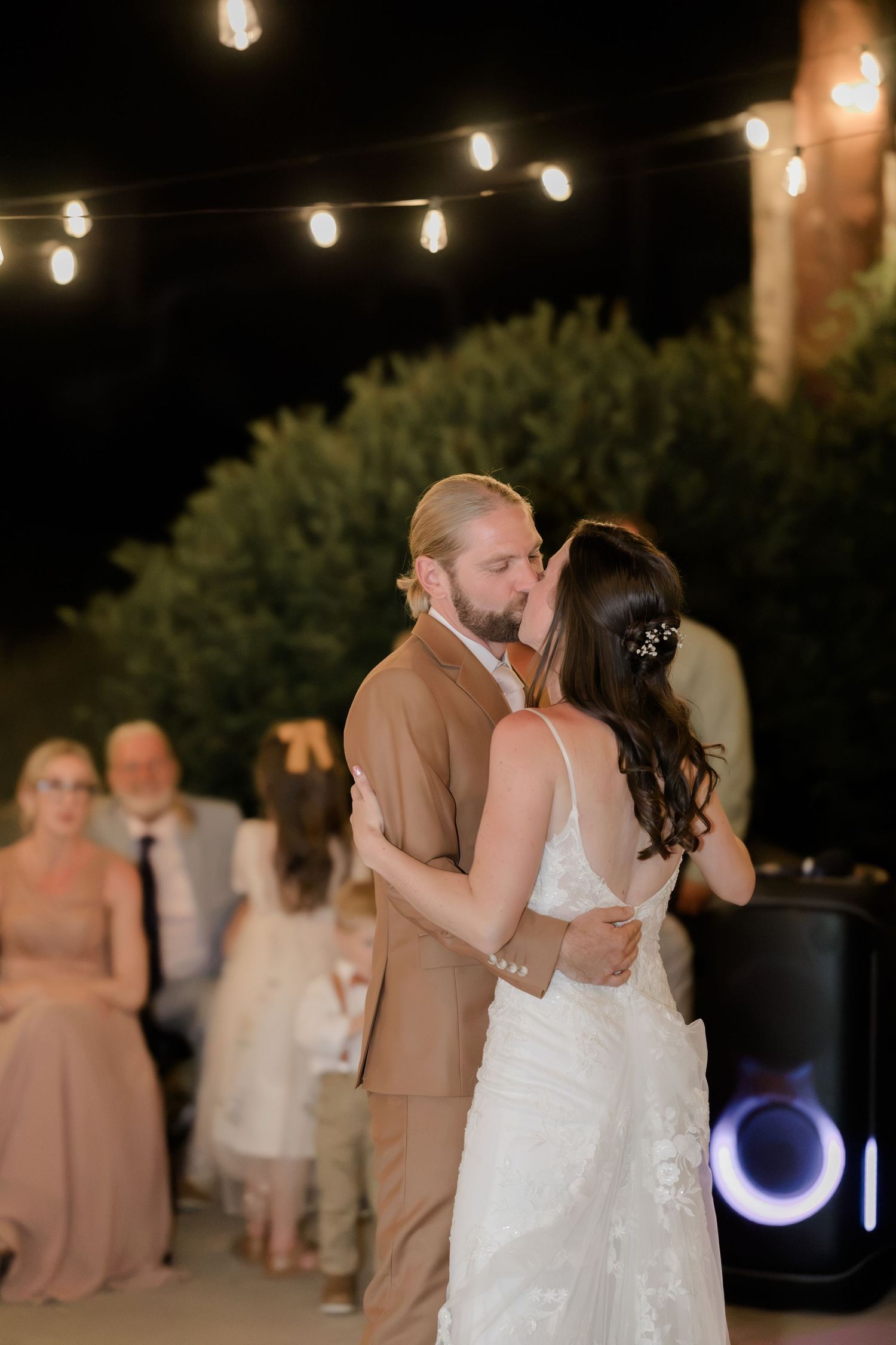 Bride and groom share a romantic first dance kiss outdoors at night under string lights at their wedding reception.