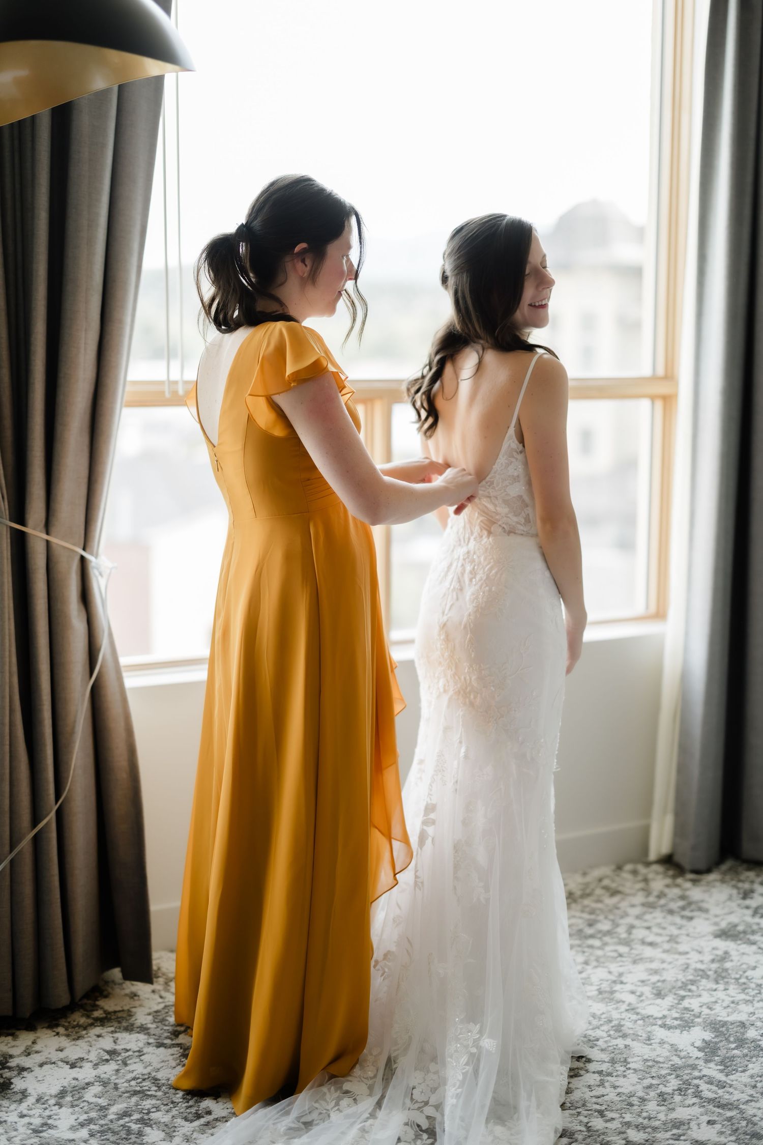 Bridesmaid in yellow gown helps bride in lace wedding dress near window in elegant hotel room.
