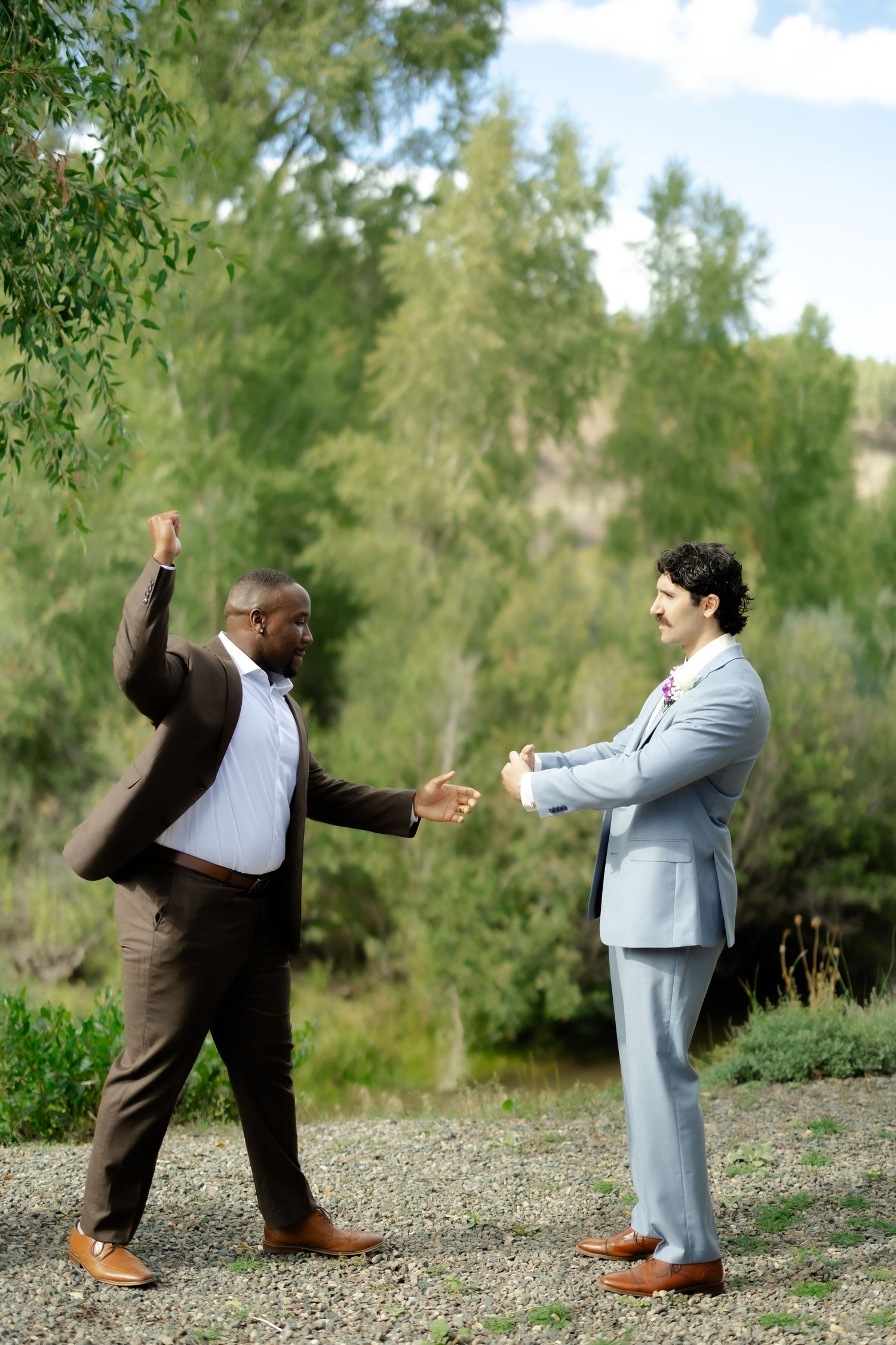 Two men in suits celebrate outdoors, one raising fist in triumph while the other extends hand for a handshake.