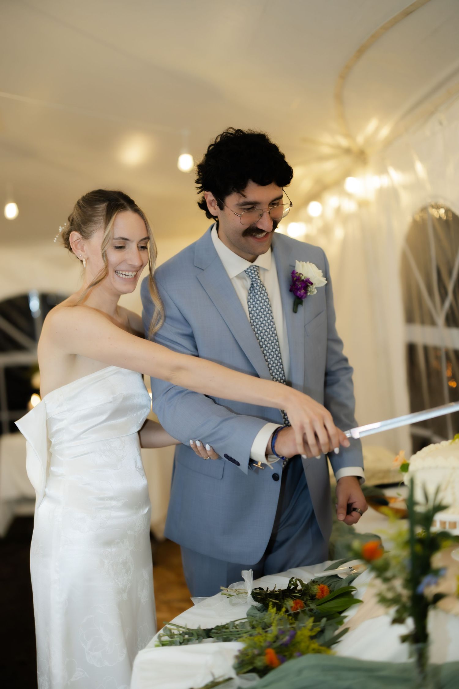 Bride and groom cutting wedding cake together under string lights at elegant tented reception venue.