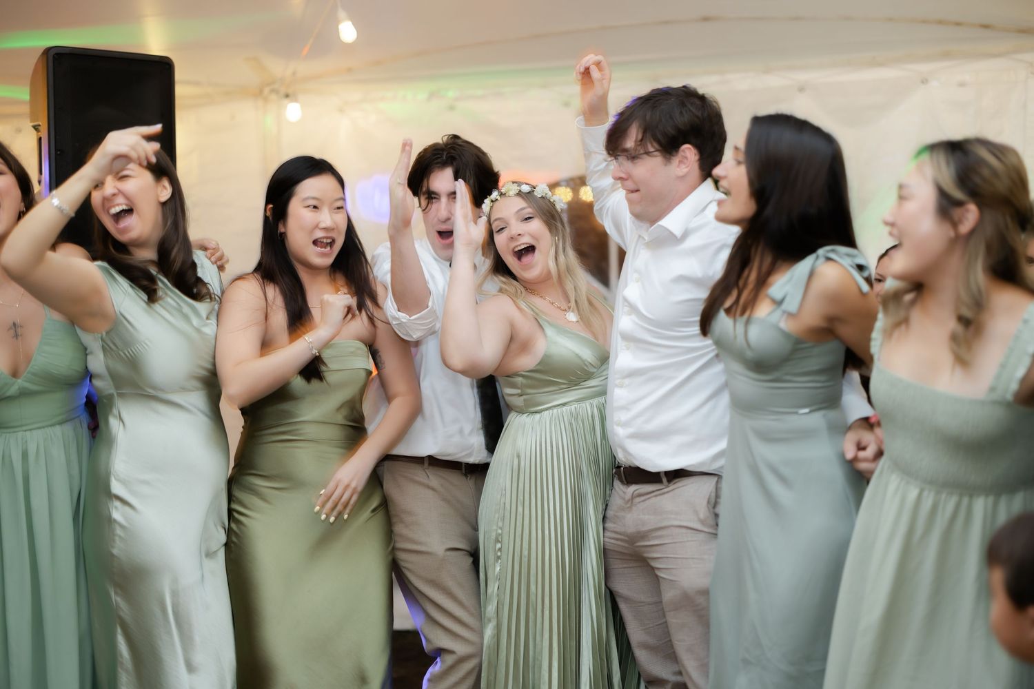 Bridesmaids in sage green dresses dancing and laughing with guests at a lively wedding reception.