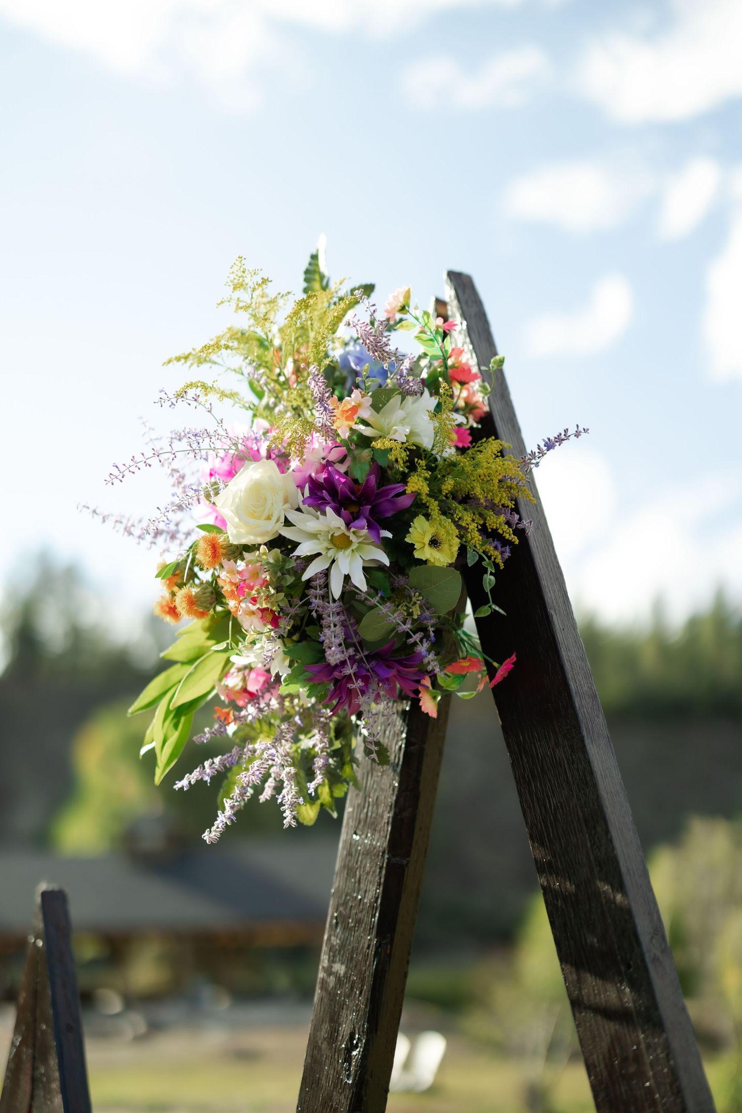 Colorful wildflower bouquet arranged on a rustic wooden ladder arch outdoors under a bright blue sky.