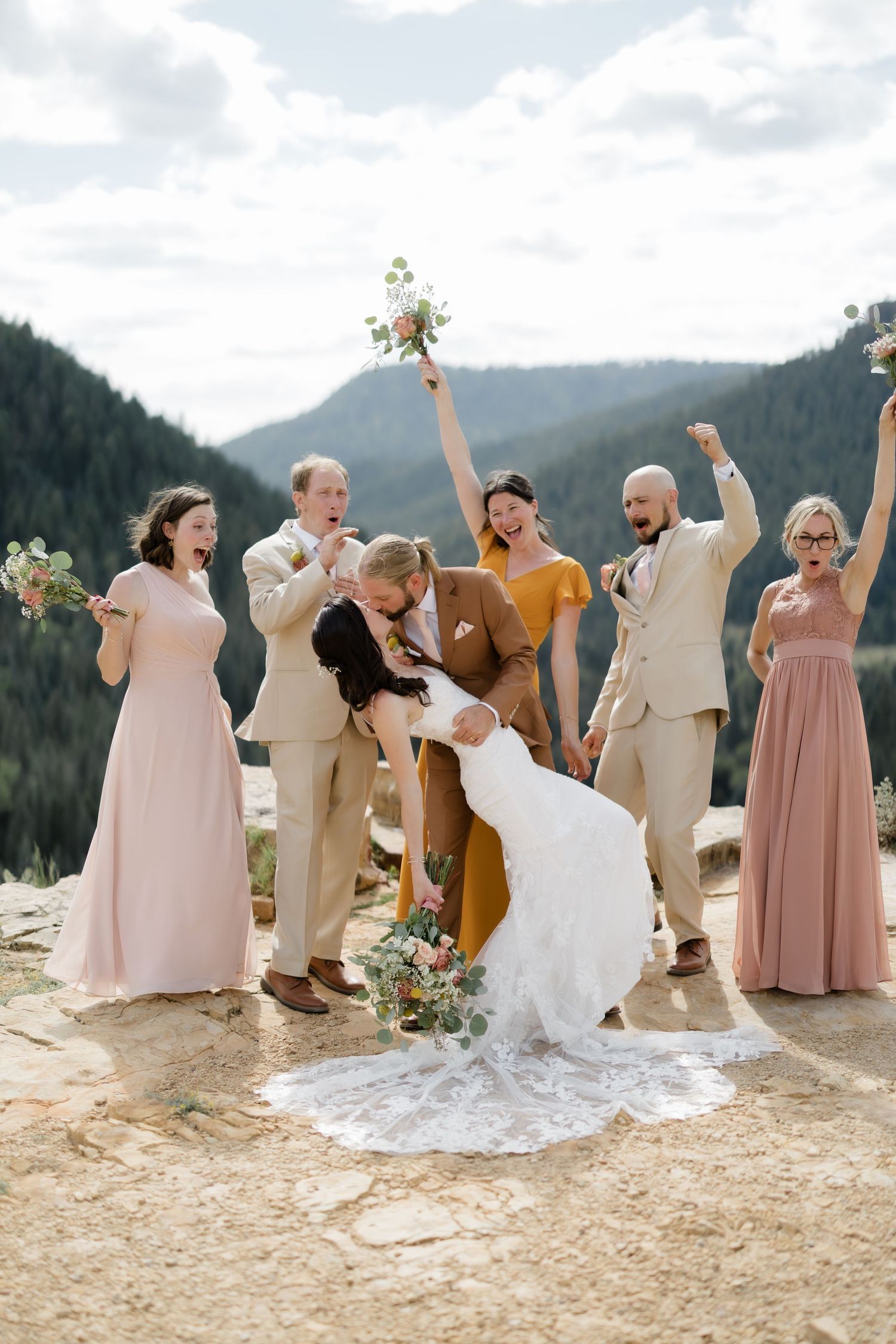 A joyful wedding party celebrates as the bride and groom share a dip kiss on a rocky mountain overlook.