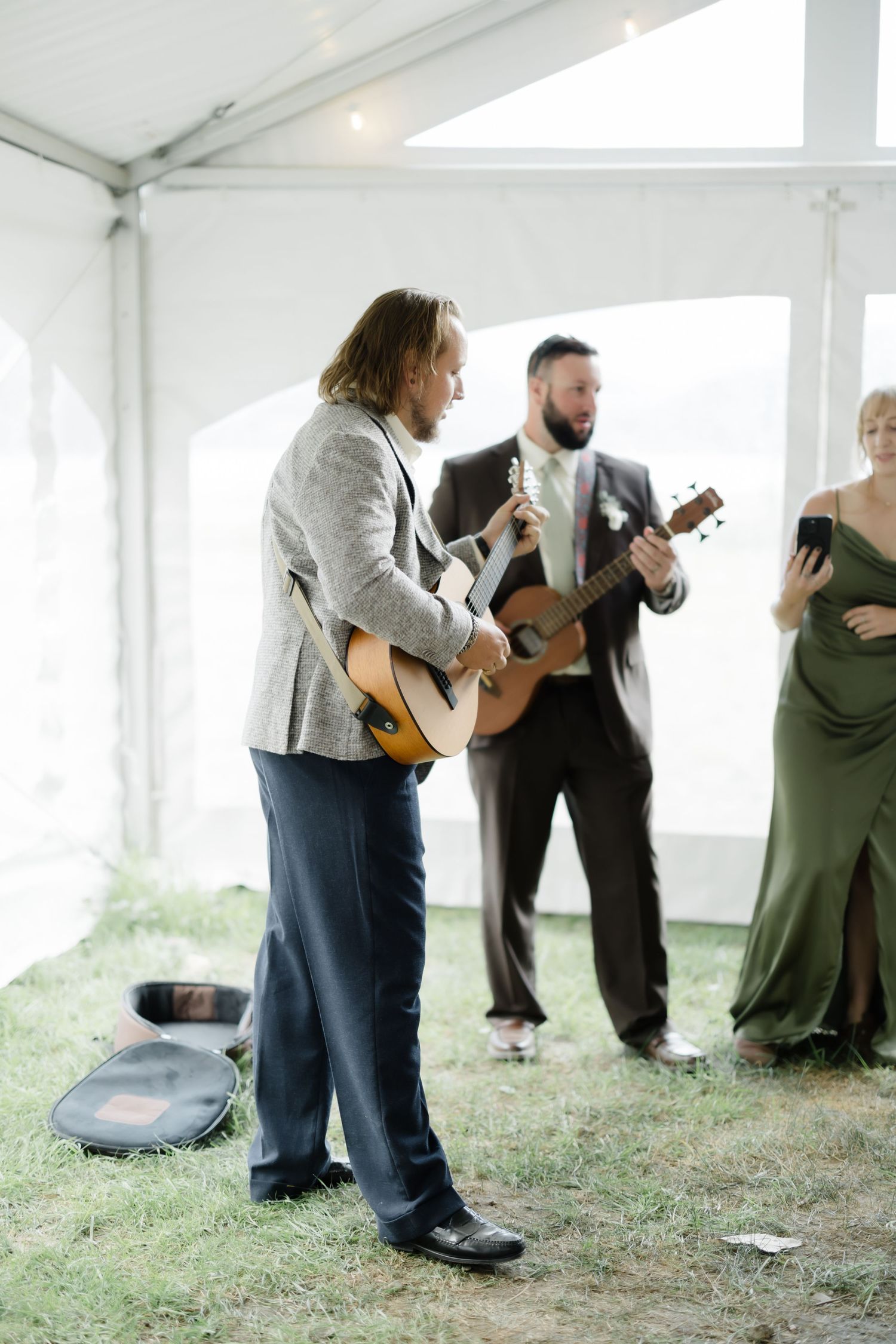 A musician plays acoustic guitar at an outdoor wedding reception under a white tent.