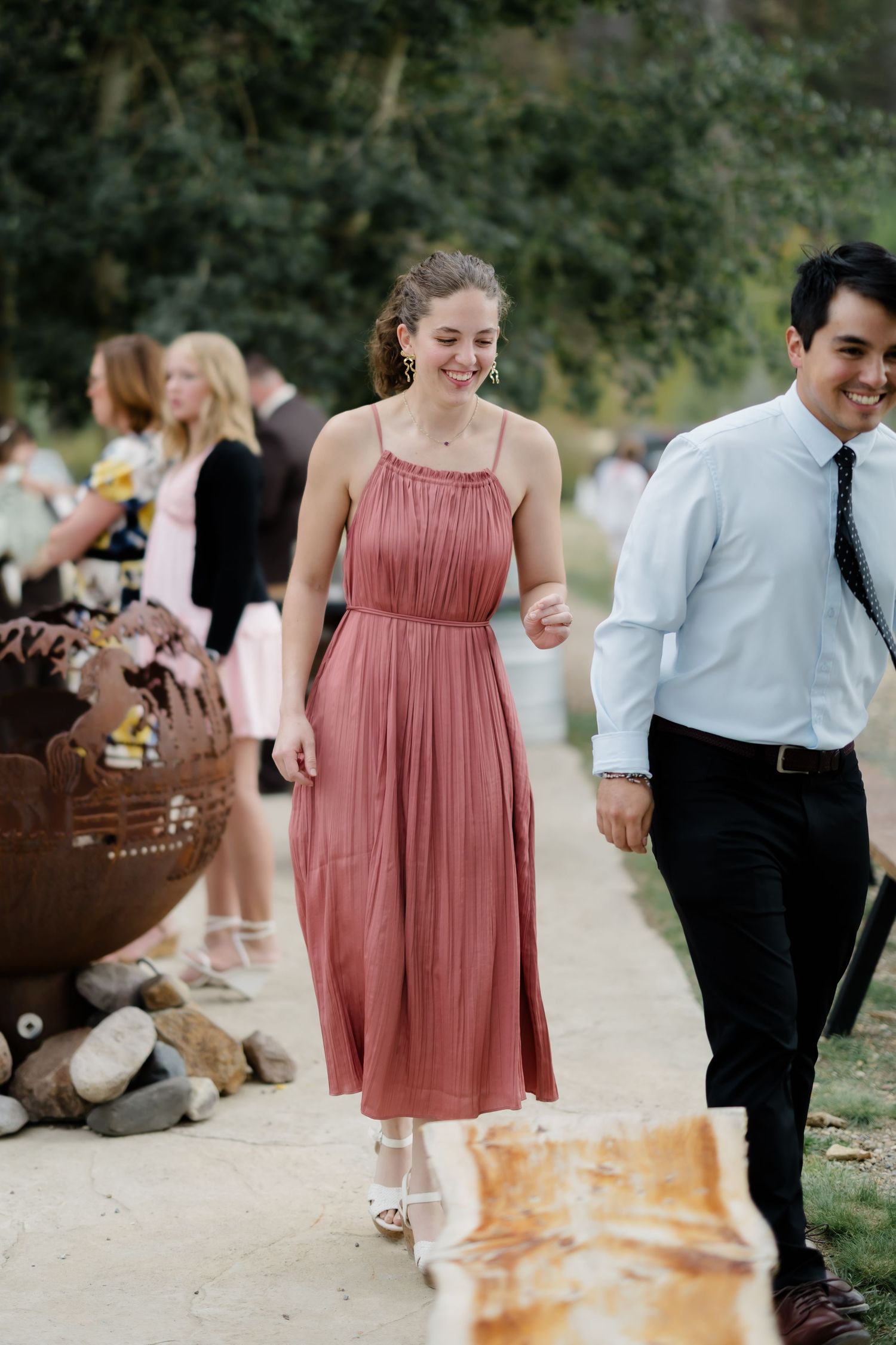 Woman in pink midi dress smiling at outdoor wedding reception with guests and fire pit in background.