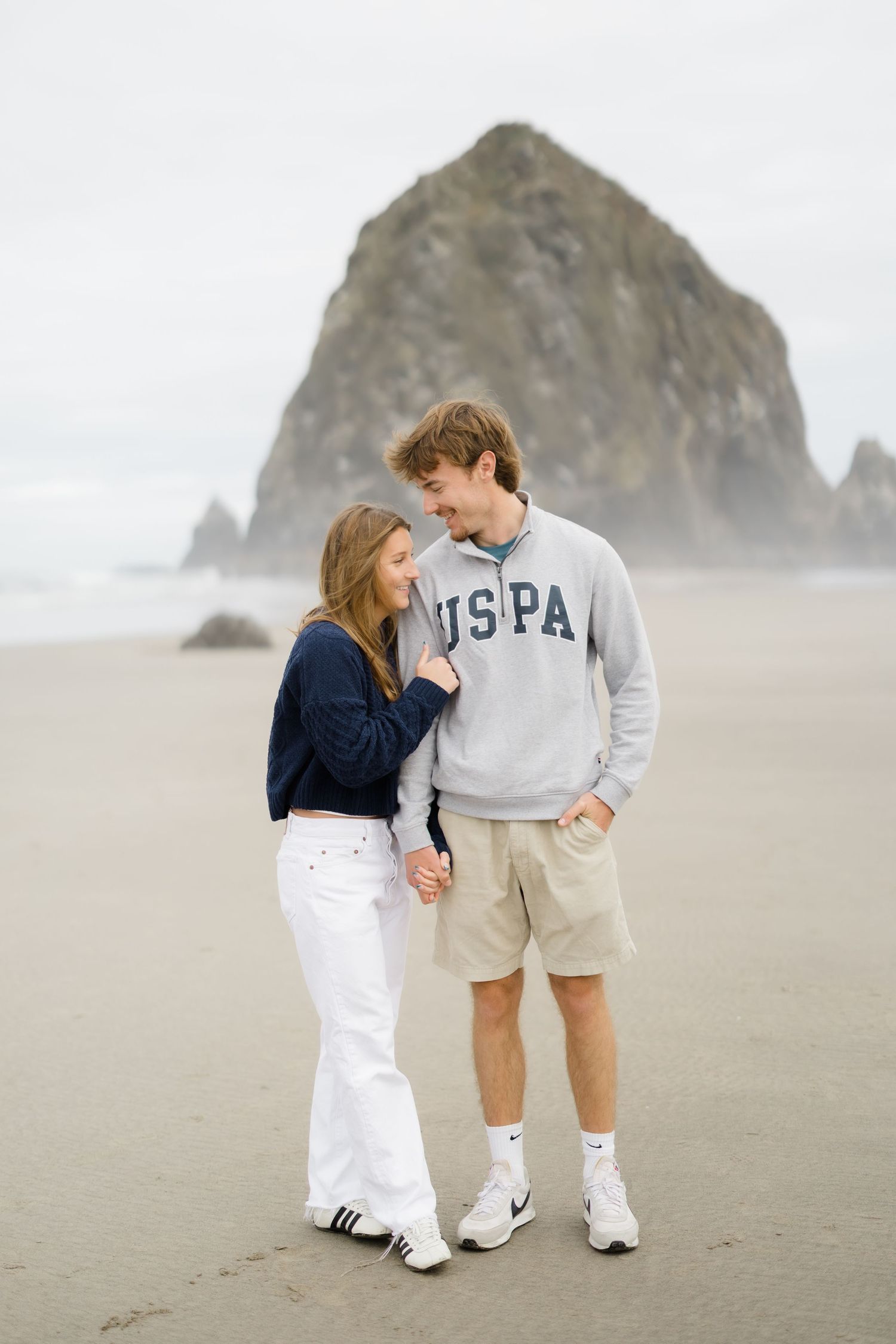 Couple holding hands and smiling on a beach with Haystack Rock in background, woman in navy top and white pants.