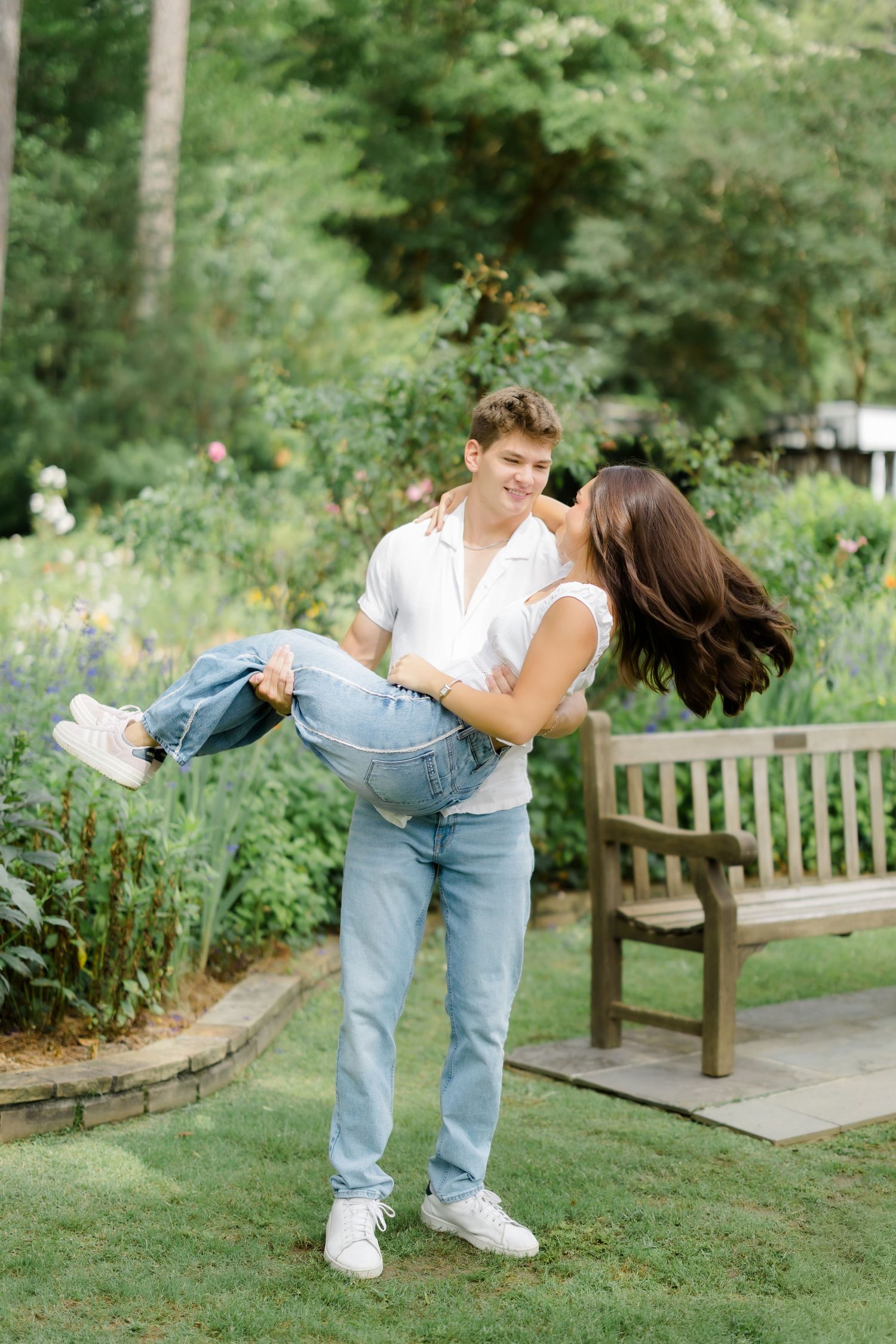 A young man carries a woman in his arms in a lush garden, both smiling and wearing jeans and white tops.