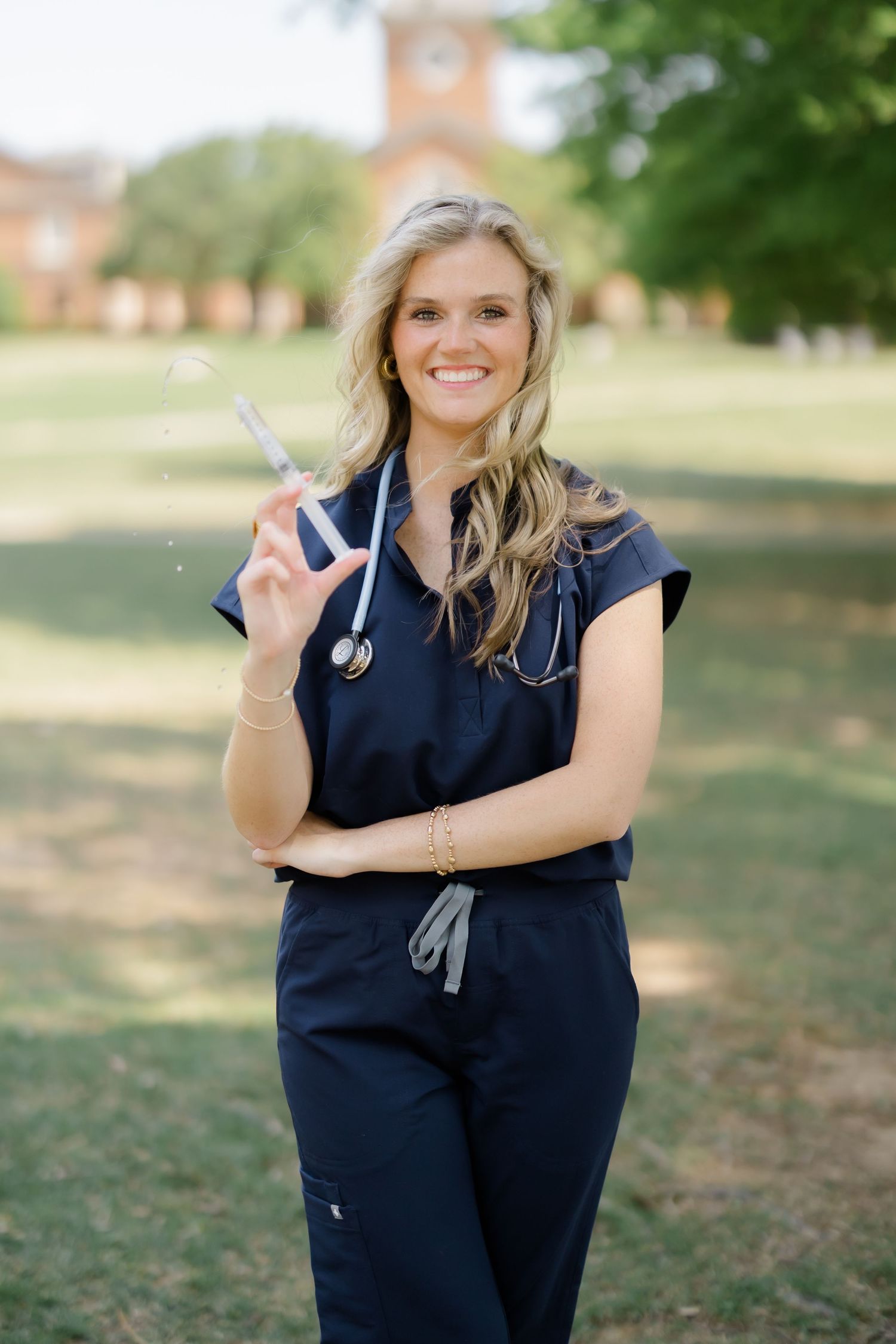 Smiling female nurse in navy scrubs with stethoscope holding medical tool outdoors on green campus grounds.