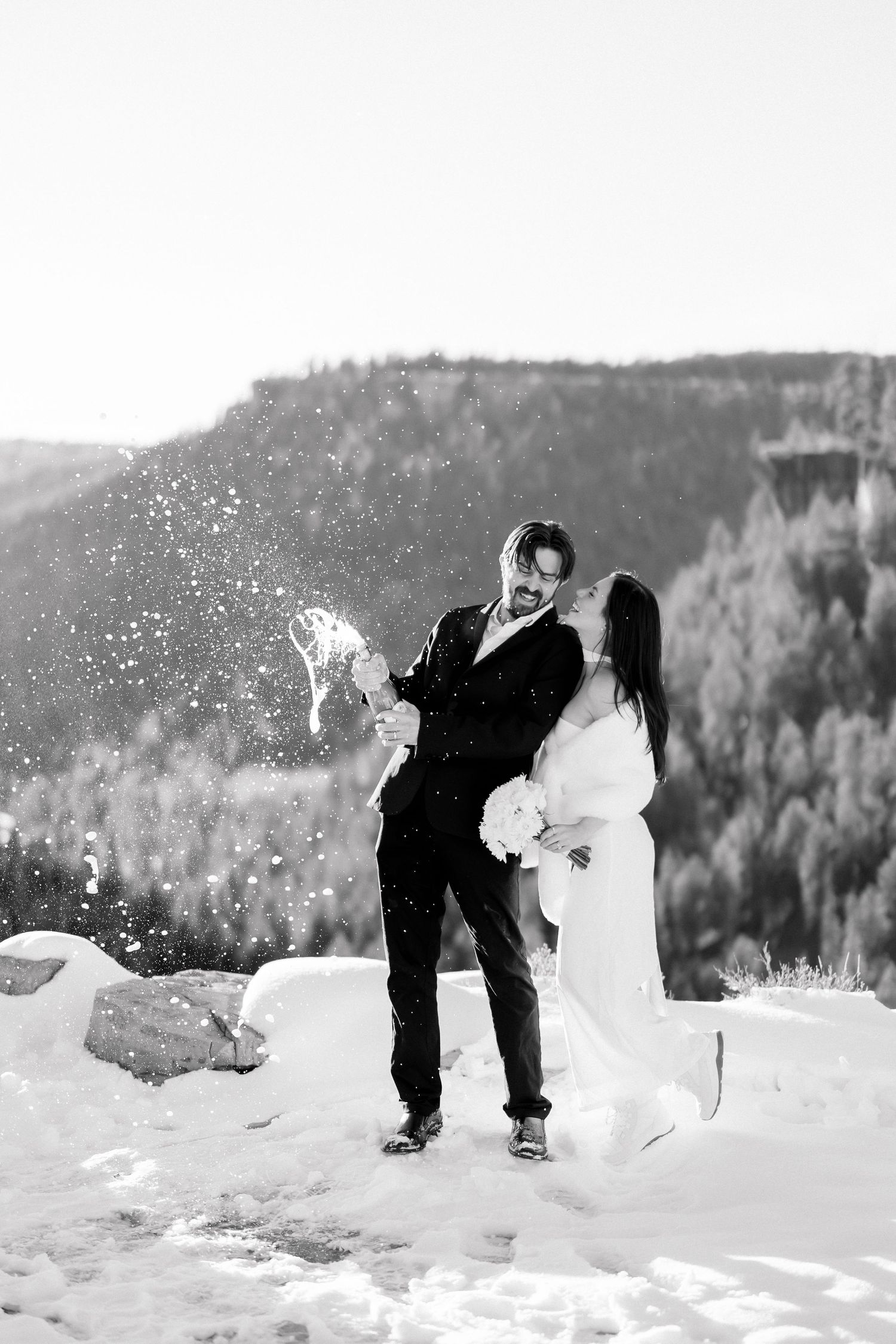 Bride and groom celebrate in snowy mountain landscape, groom tosses snow joyfully, black and white wedding photo.