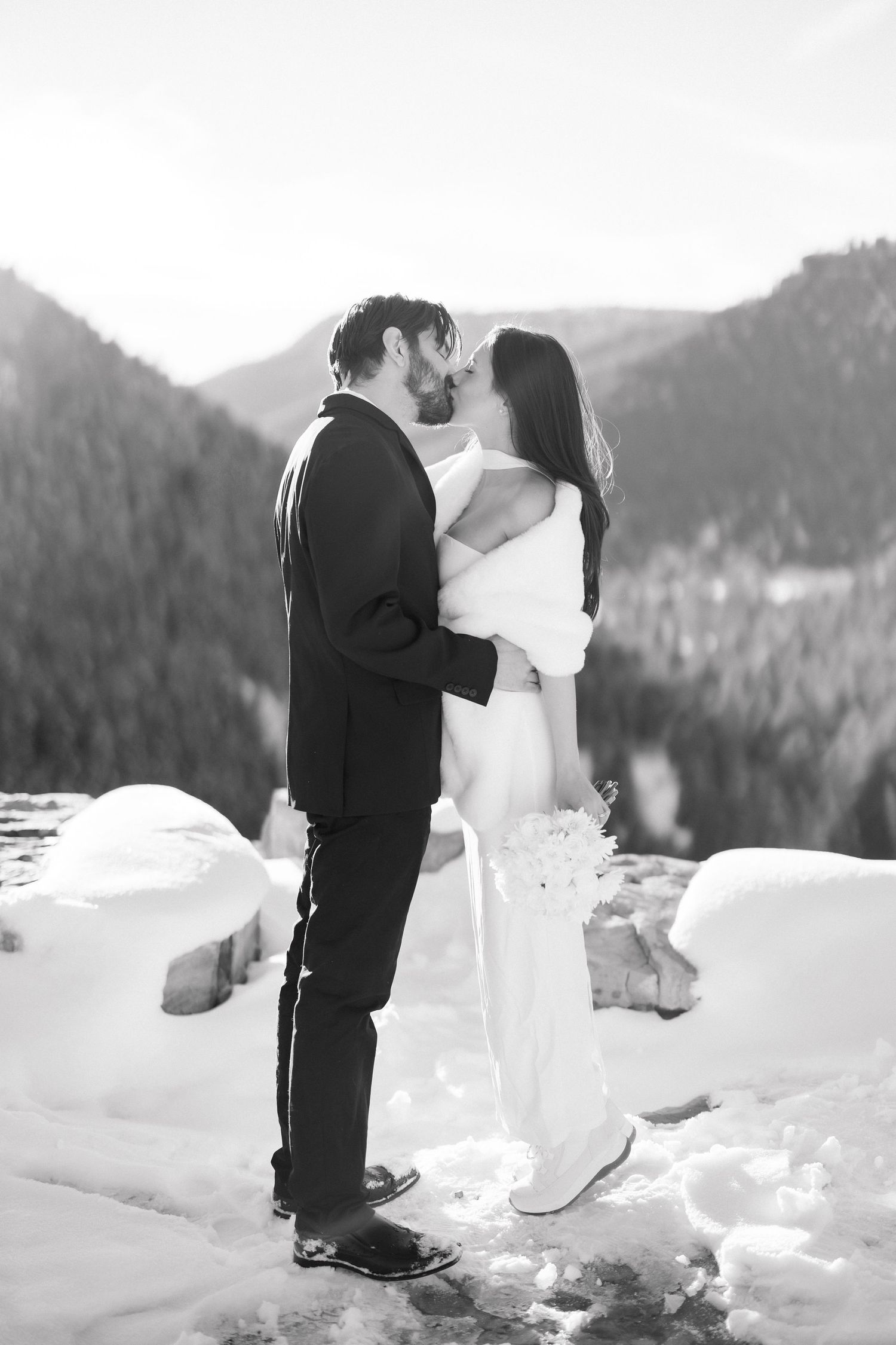 Black and white image of bride and groom kissing in snow with mountain forest behind them.