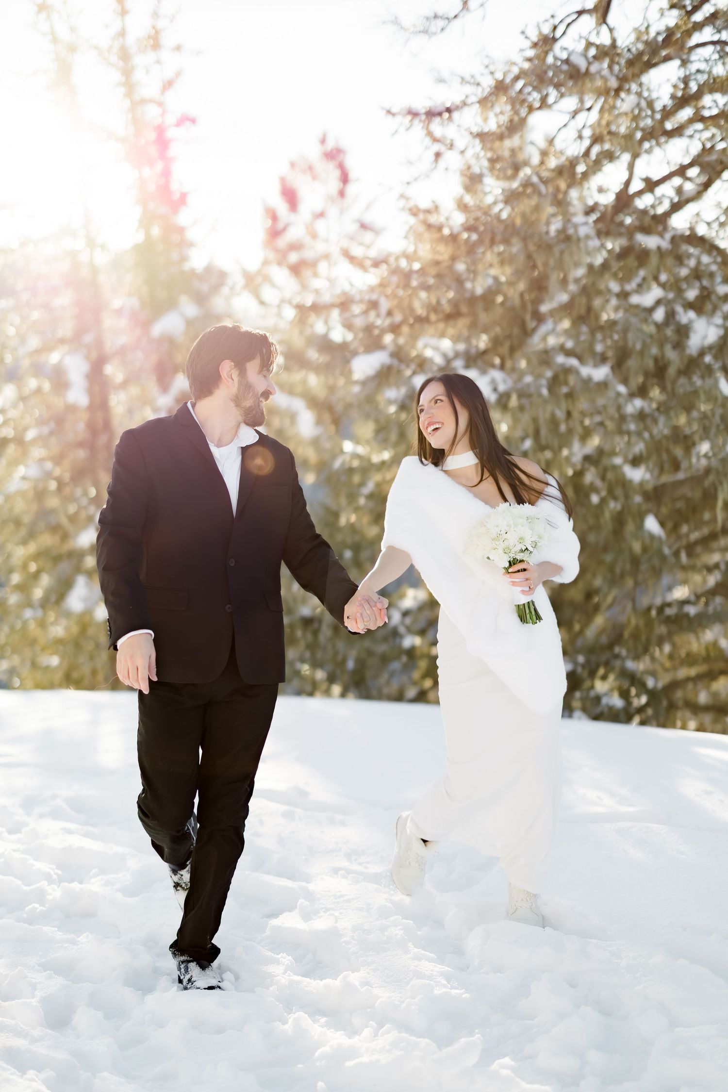 Joyful bride and groom walk hand in hand through snow with sunlit pine trees behind them.