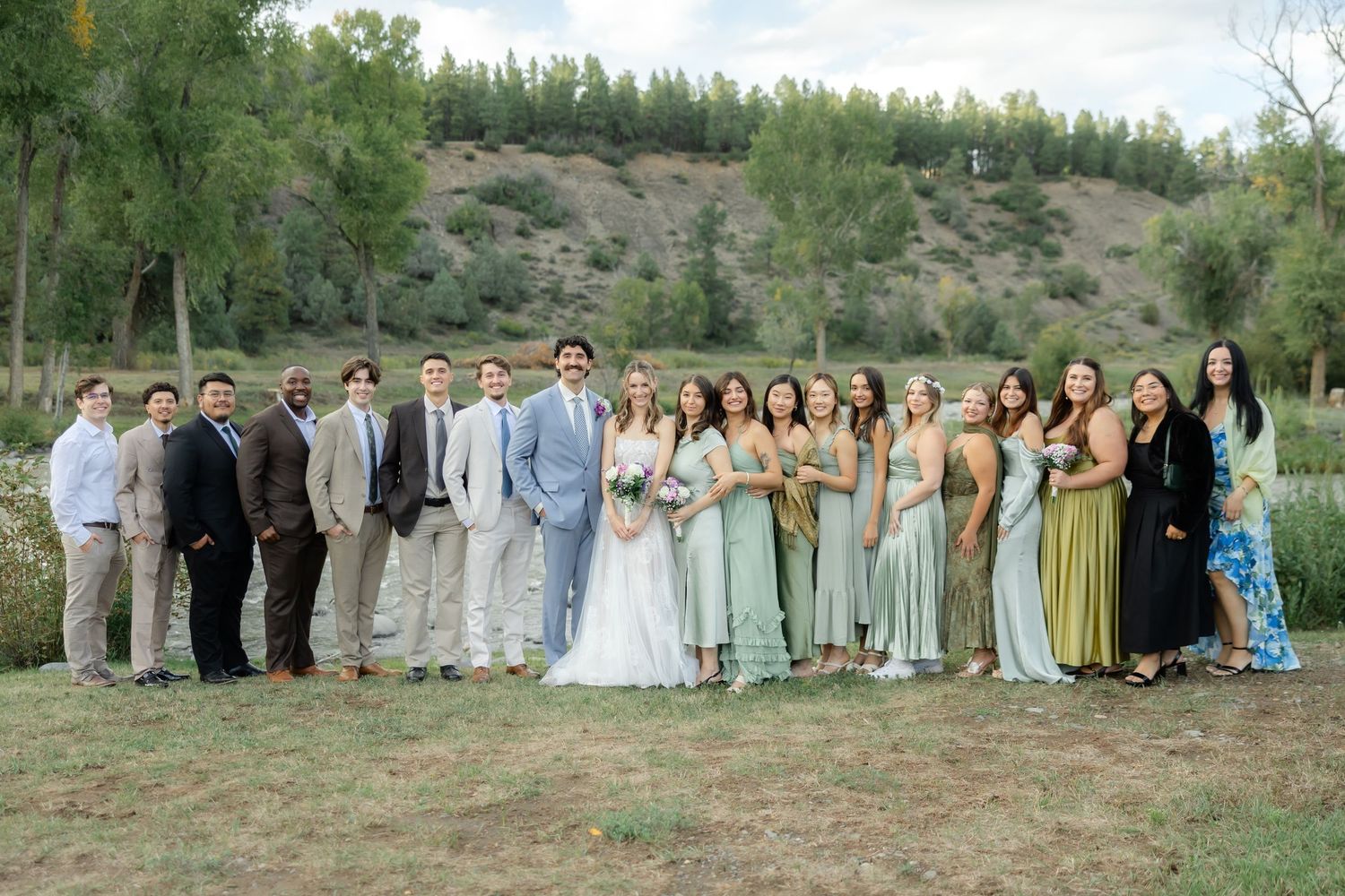 Wedding party posing outdoors in nature, bridesmaids in sage green dresses, groomsmen in tan suits, bride and groom centered.