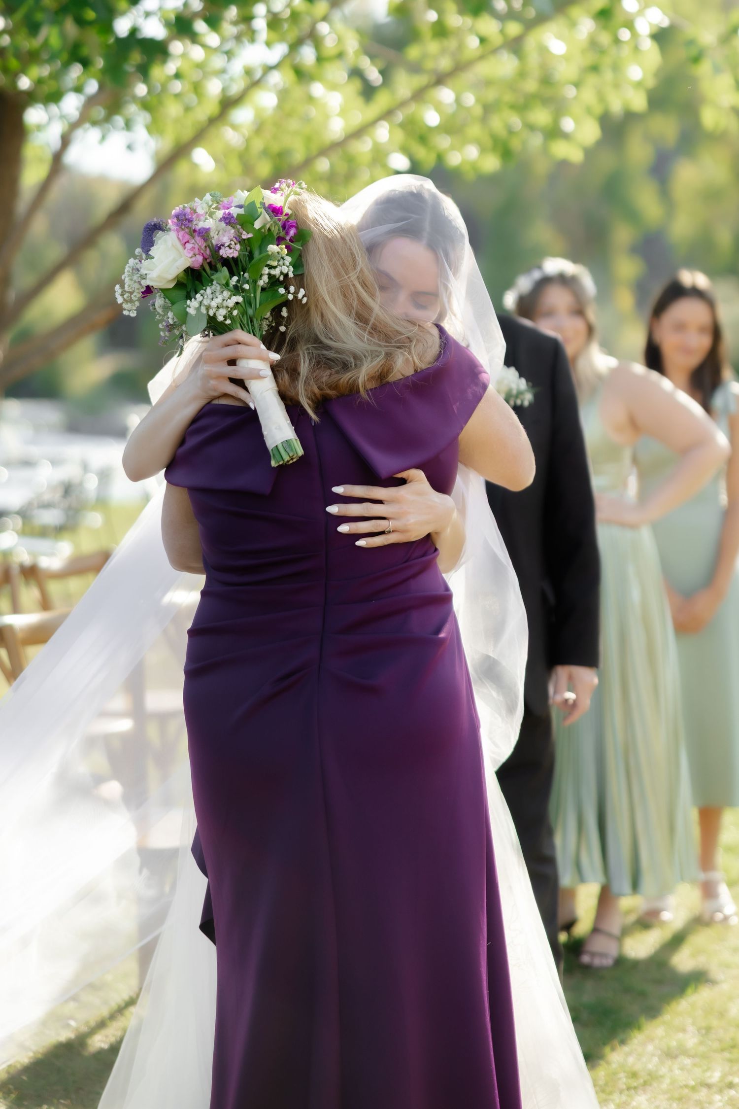 Bride in white gown and veil hugging woman in purple dress at outdoor wedding ceremony.