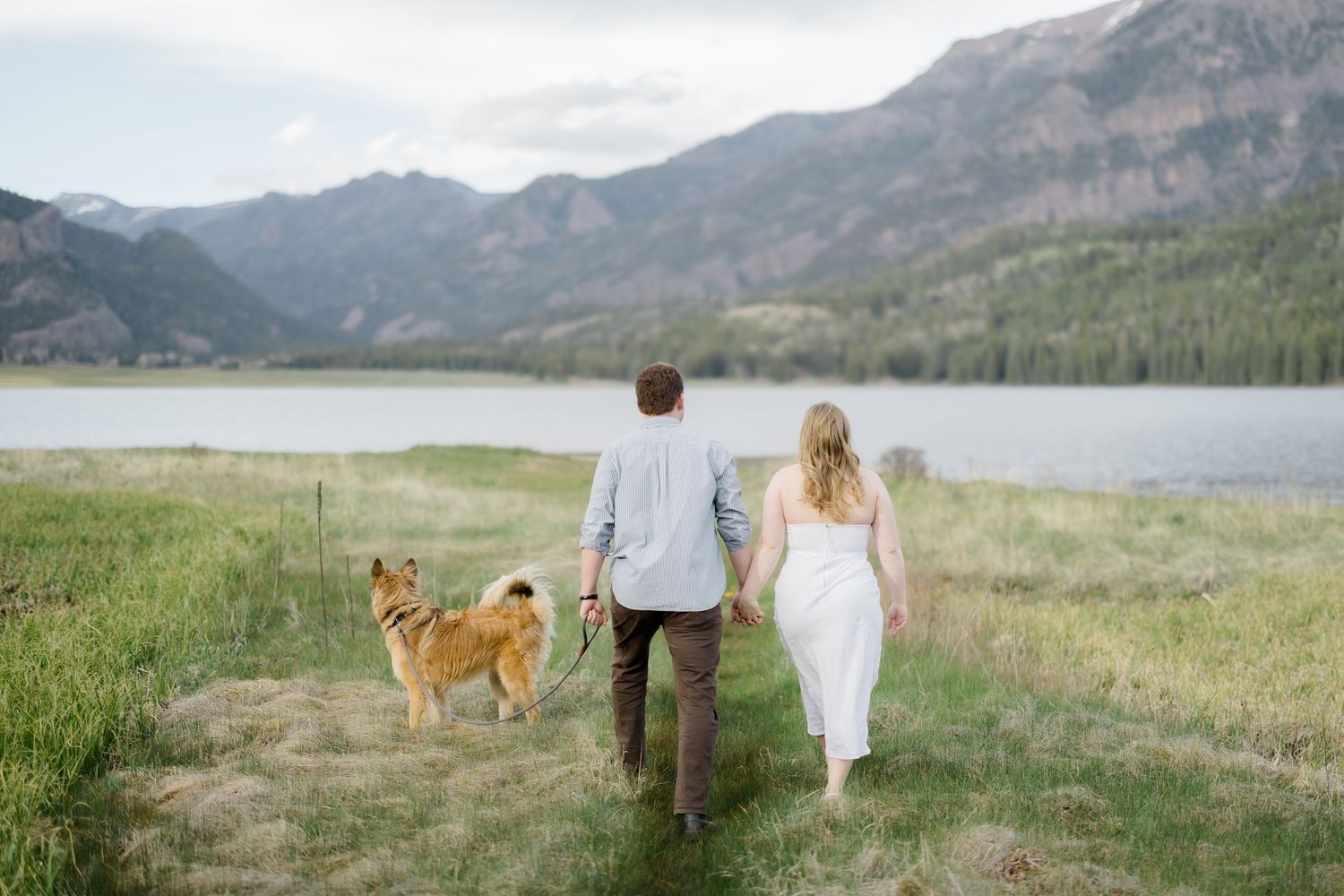 Couple walking hand-in-hand with their golden dog toward a mountain lake, surrounded by lush green meadows.