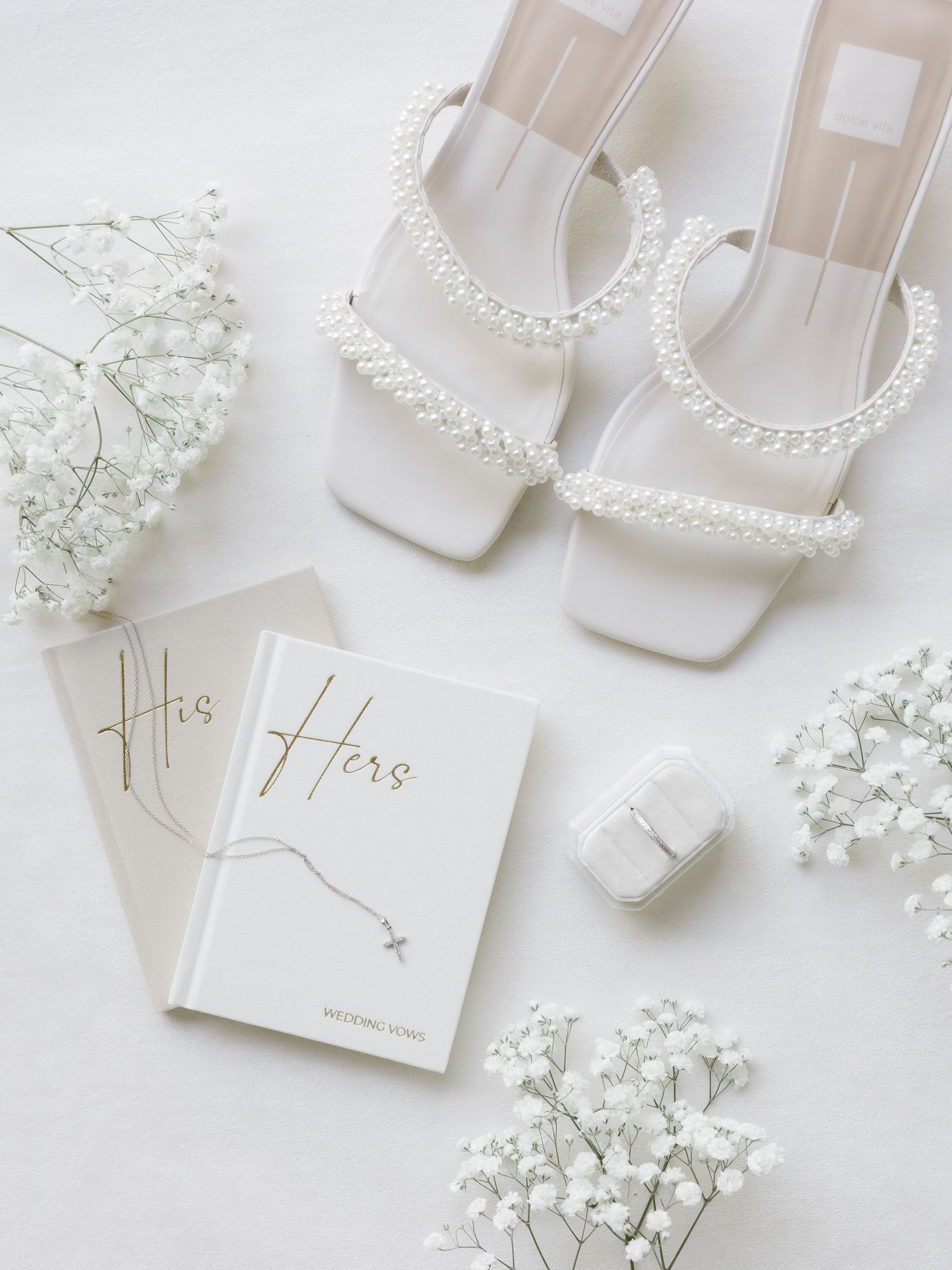 White pearl-strapped bridal heels beside His and Hers vow books, baby's breath flowers, and a ring box.